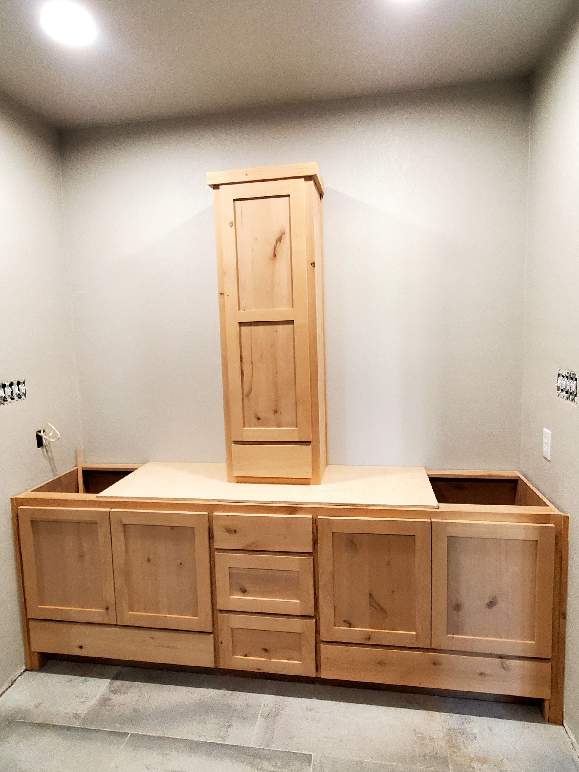 Light wood bathroom cabinetry, including base cabinets and tall cabinet, unfinished against a neutral wall.