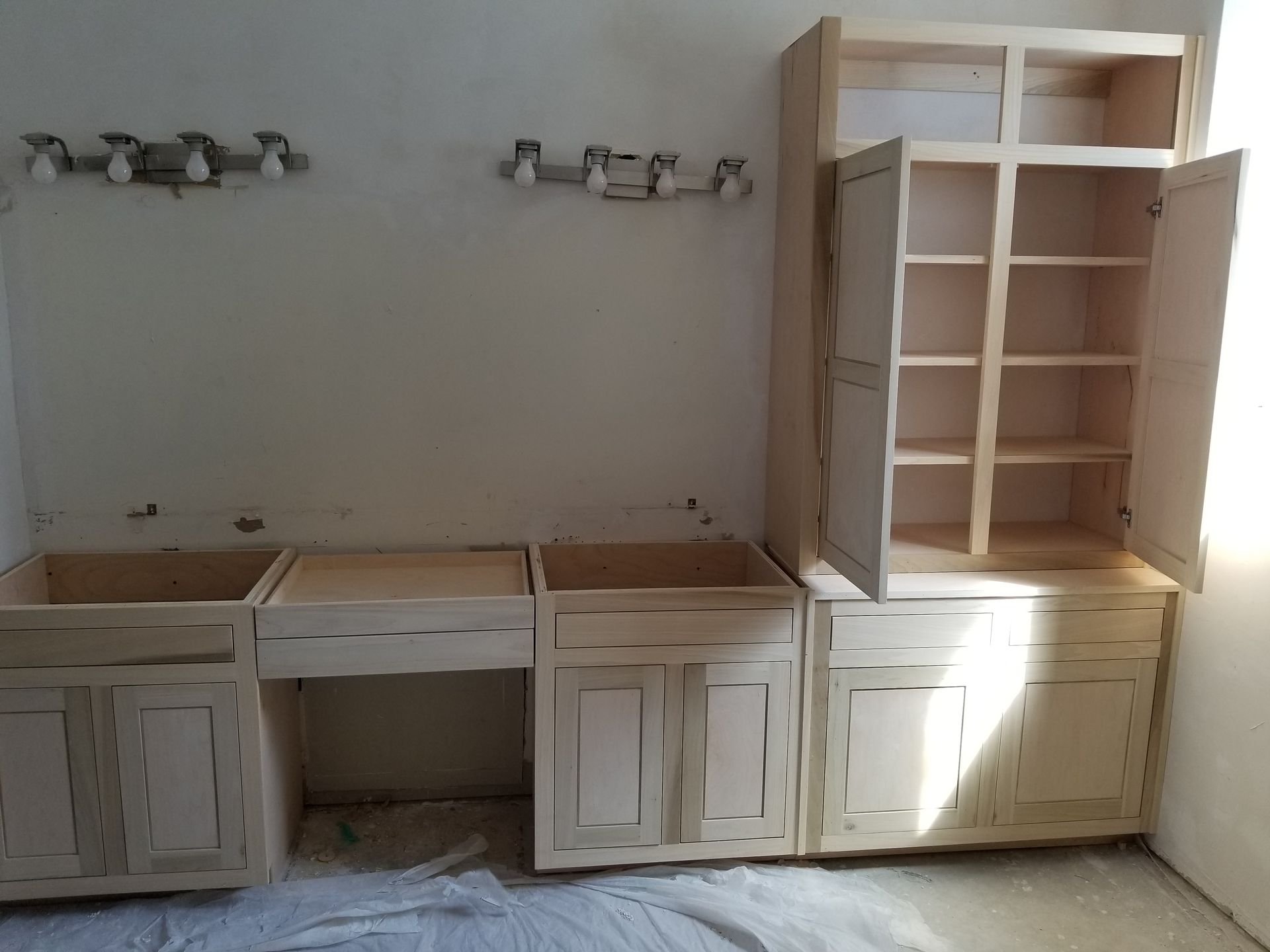 Unfinished kitchen cabinets installed against a wall, with open doors and shelving. Two light fixtures are above.