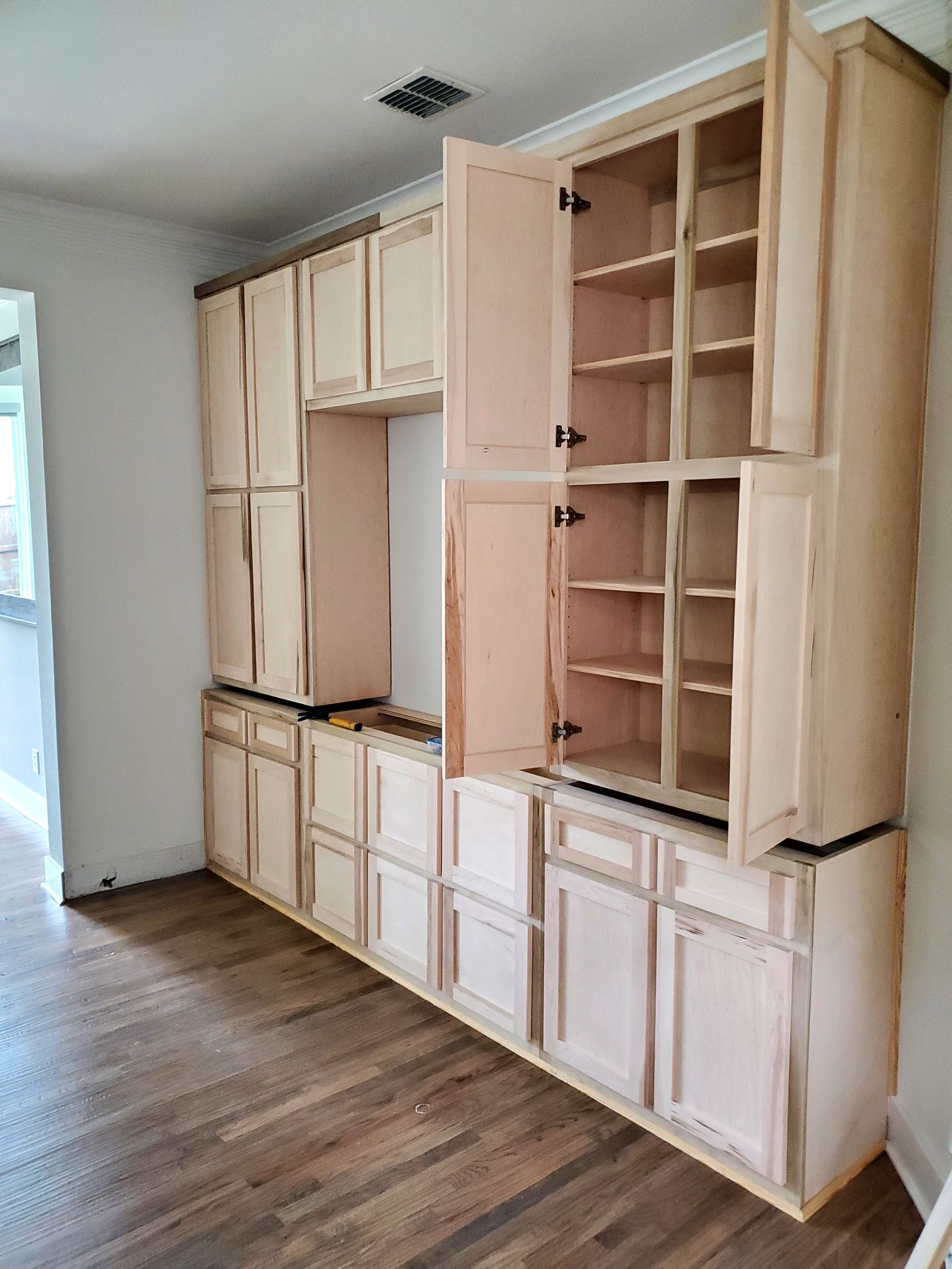 Unfinished light wood kitchen cabinets being installed in a room with hardwood floors.