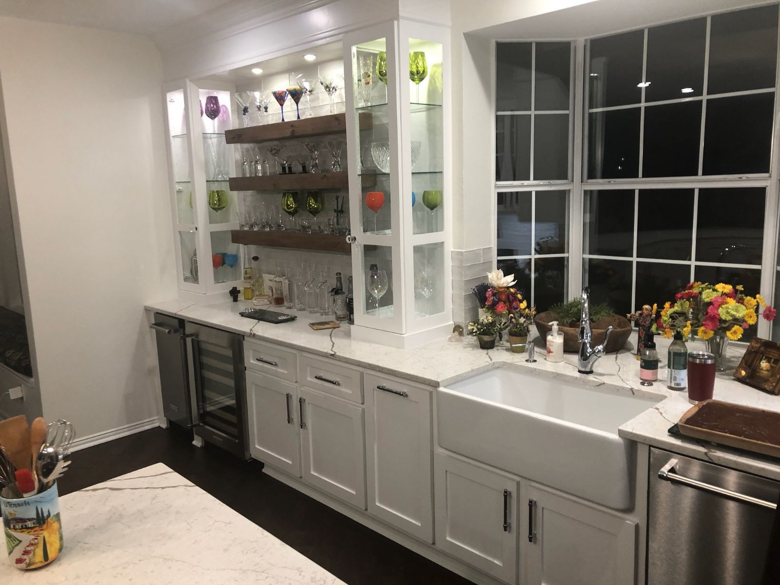 White kitchen with built-in glass cabinets, wine fridge, and farmhouse sink by a large window.