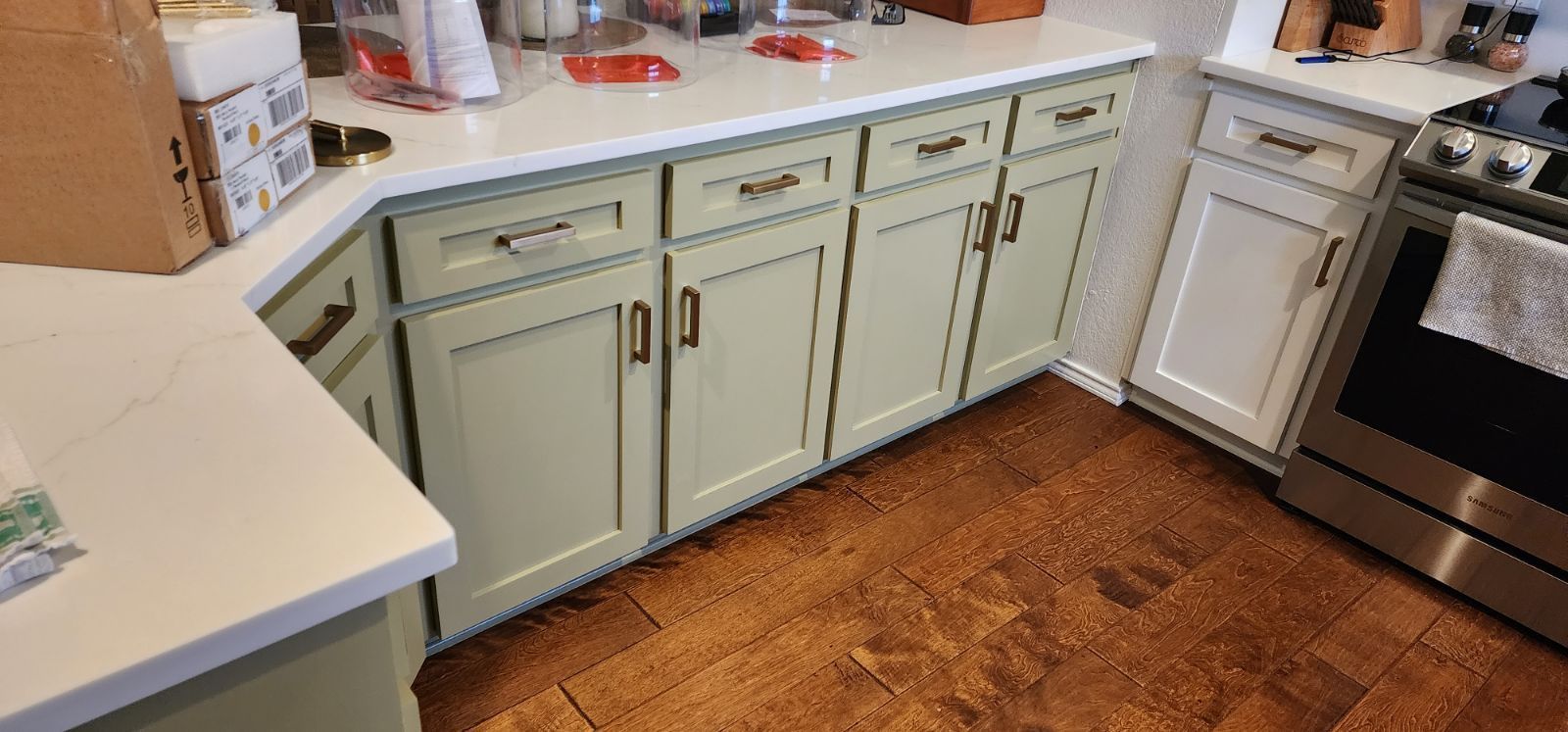 Kitchen cabinets painted a pale green color with dark hardware, set against a white countertop and brown hardwood floor.