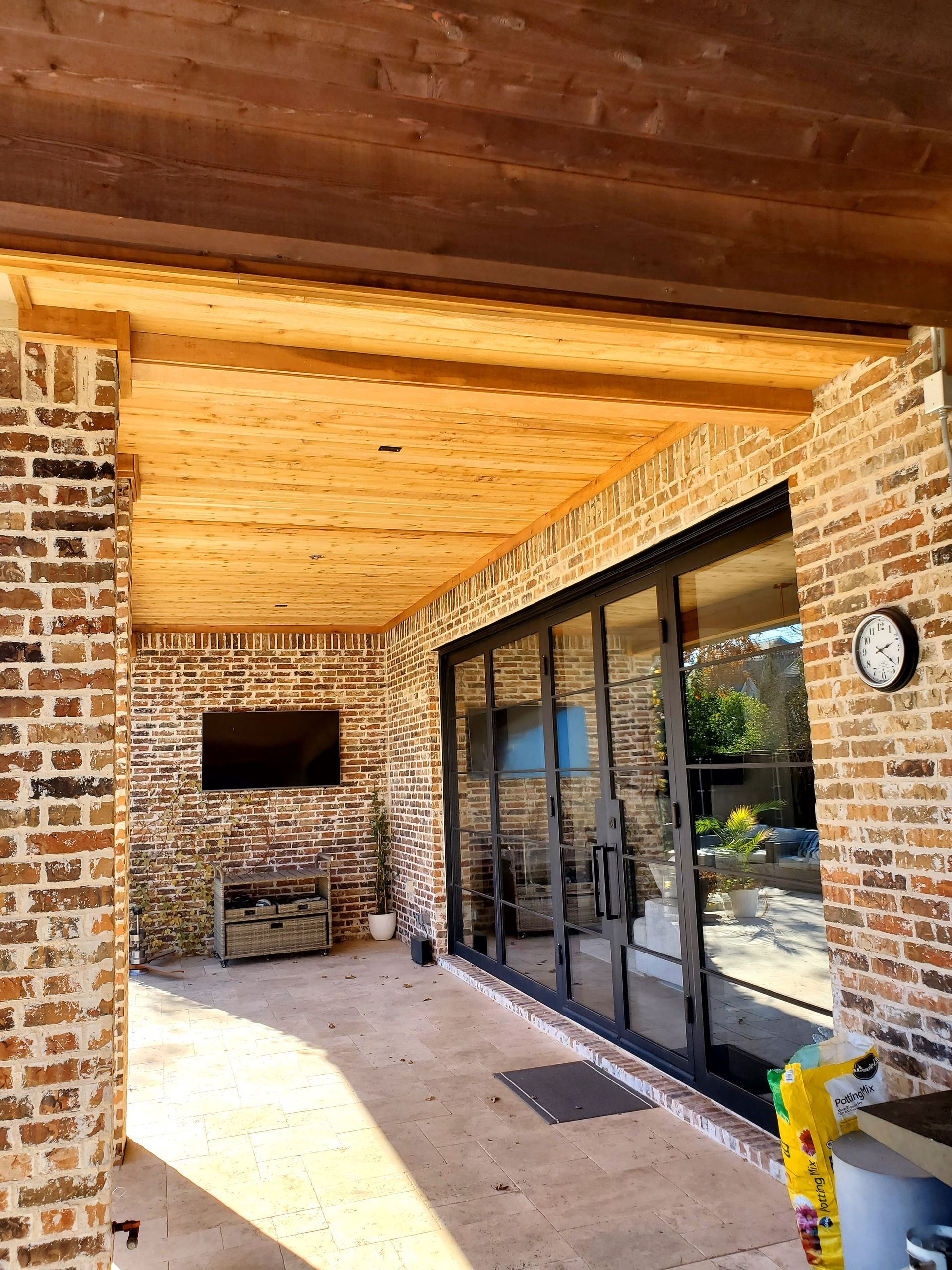 Covered outdoor patio with brick walls, wooden ceiling, and glass doors overlooking a yard.
