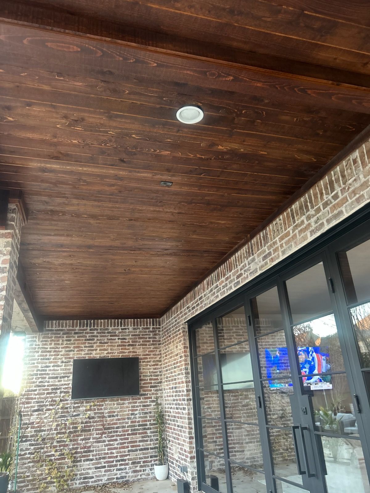 Brown wooden ceiling with recessed light, brick wall, and glass doors. A TV is mounted on the wall.