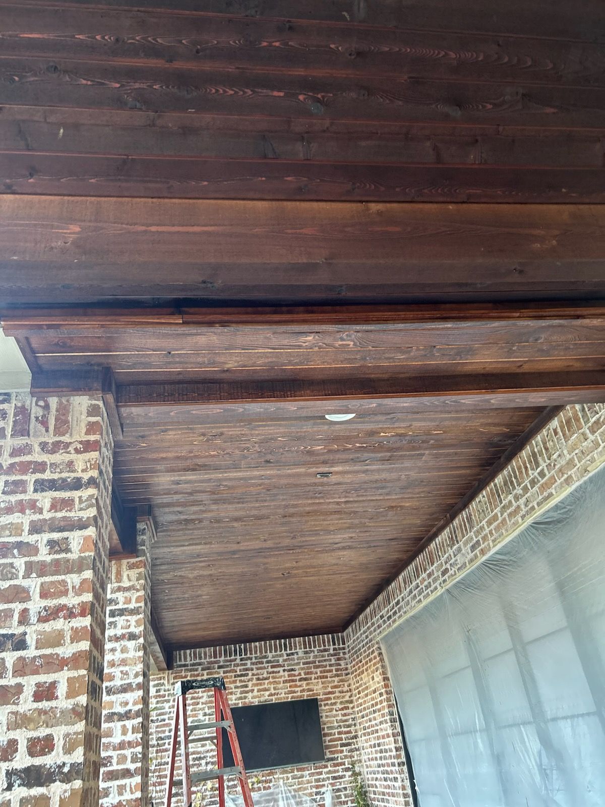 Wooden ceiling with dark stain, between brick columns and a wall. Ladder visible.