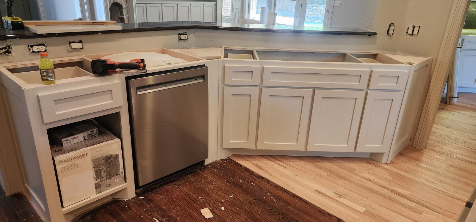 Kitchen under construction with white cabinets, stainless steel dishwasher, and wooden floor.