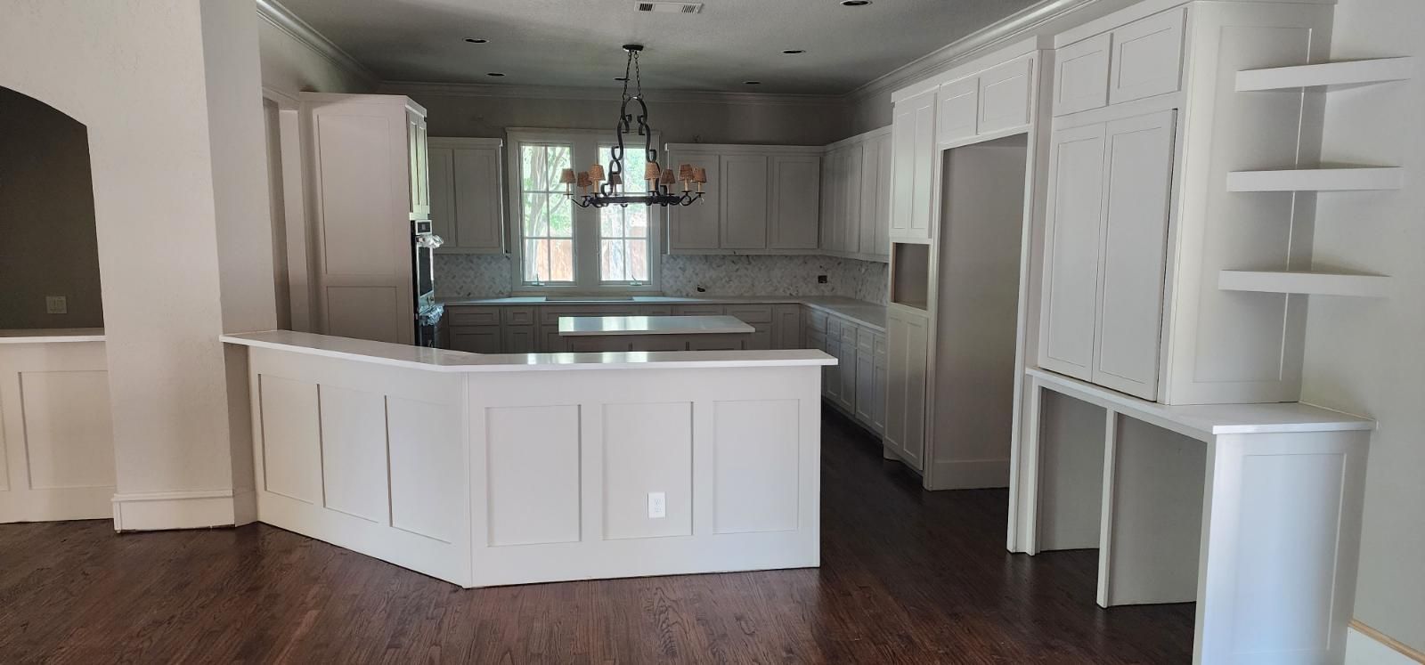 White kitchen with island, cabinets, and appliances. Dark wood floor. Chandelier hangs over island.