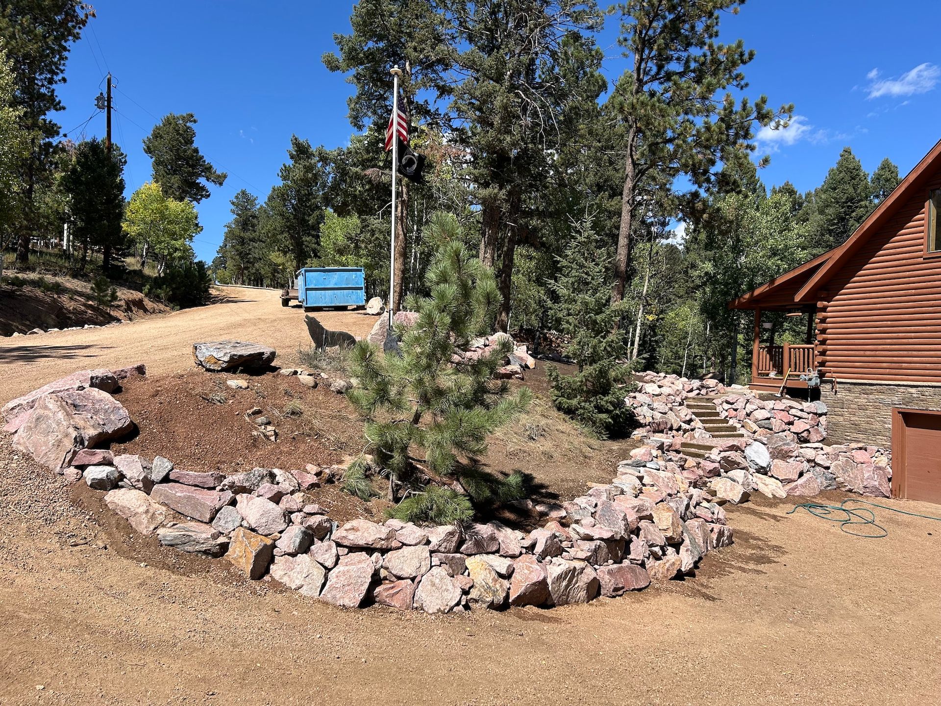 A log cabin is surrounded by trees and rocks in the middle of a dirt road.