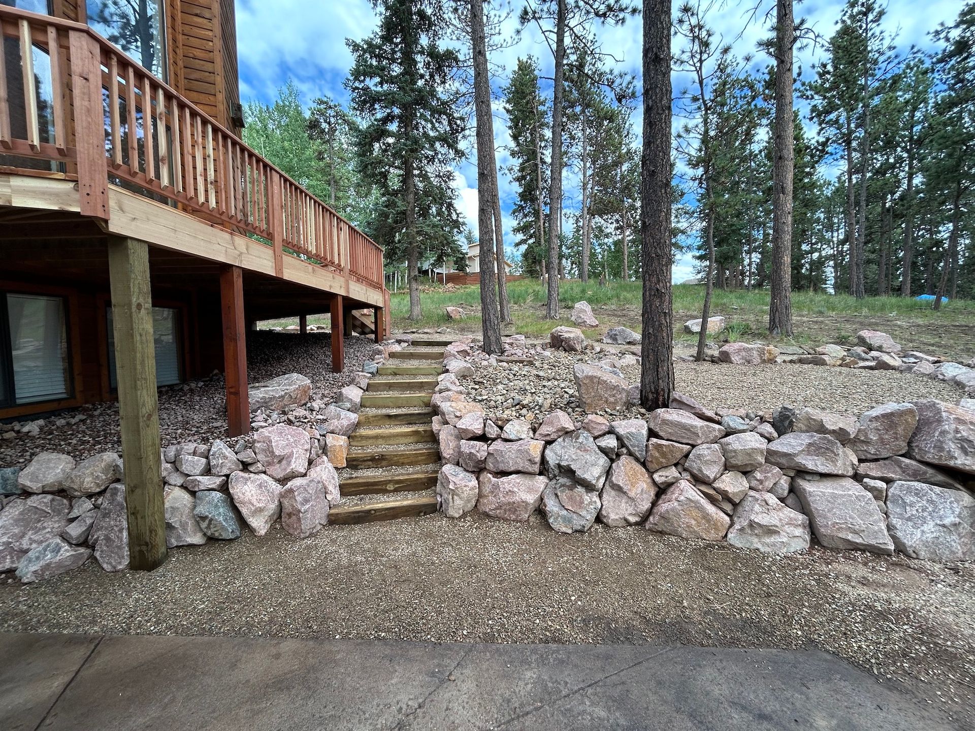 A wooden deck with stairs leading up to it is surrounded by rocks and trees.