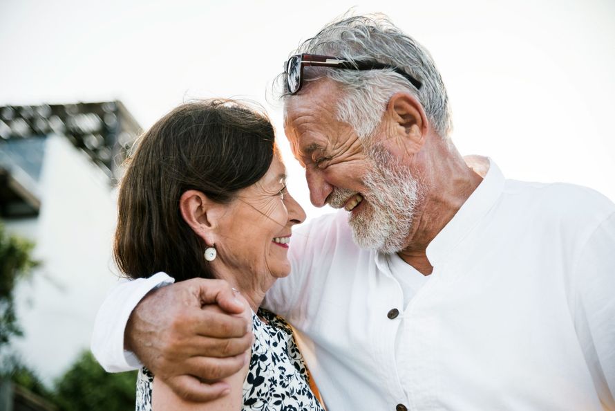 Two smiling people embrace outdoors, looking at each other warmly.
