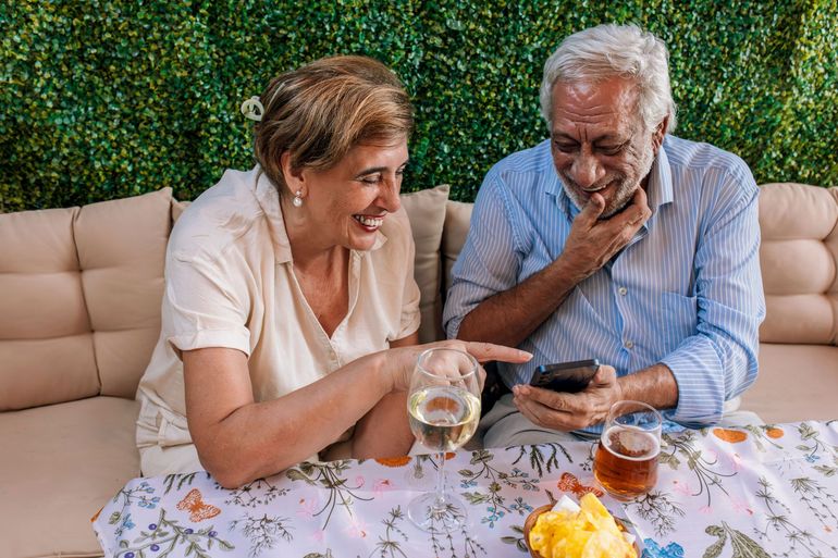 Older couple smiling and chatting at a table with tea and a phone in a cozy outdoor setting