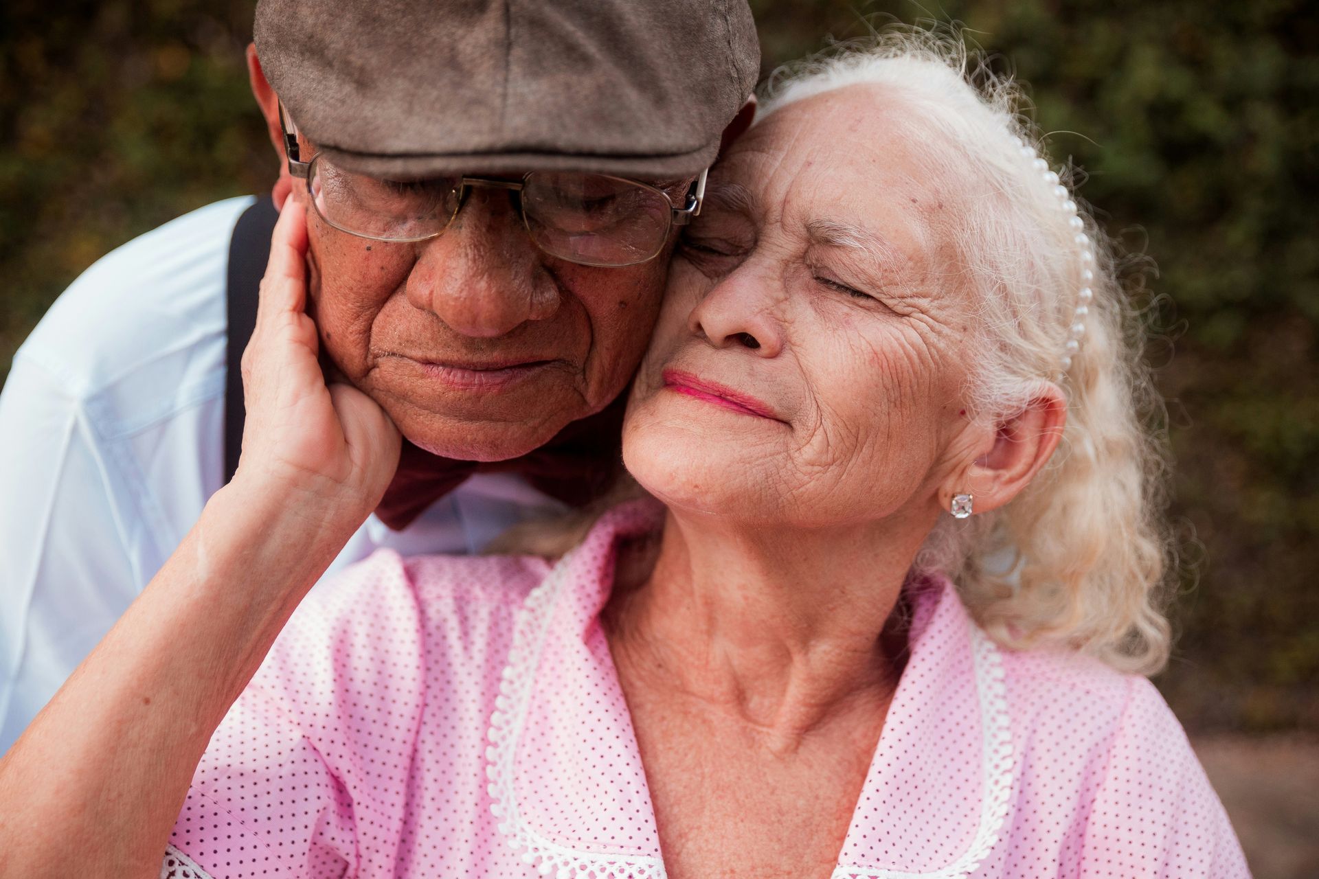 Two elderly people embracing outdoors, with one gently resting against the other and smiling