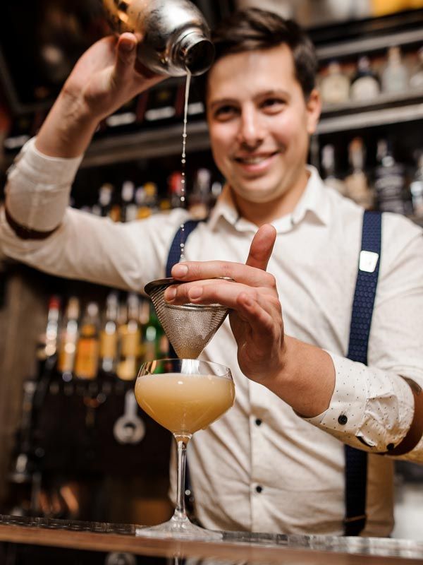 Bartender smiling while pouring a drink into a coupe glass behind a bar