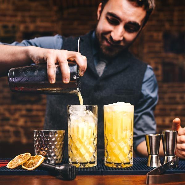 Bartender pouring a dark drink into two yellow cocktails at a bar with citrus garnish