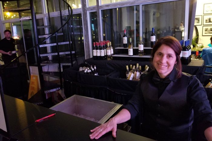 Woman smiling at a restaurant bar, with wine bottles and a staircase in the background