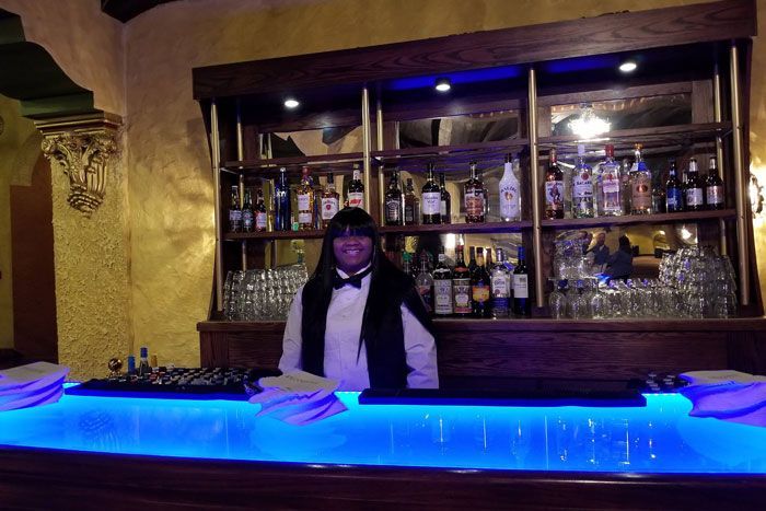 Person standing behind a blue-lit bar with bottles displayed on shelves in the background