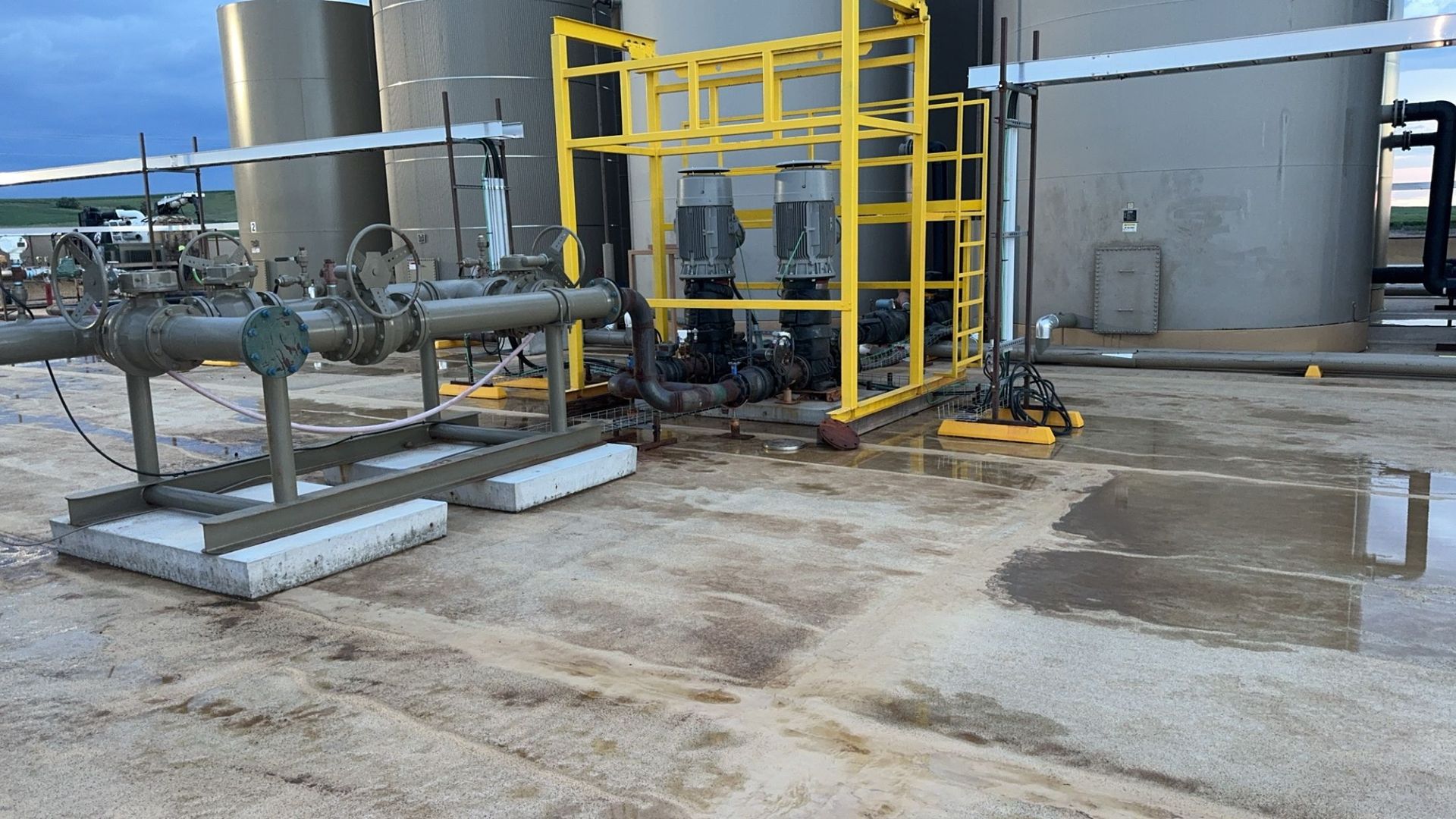 Industrial pipes and tanks on a wet concrete pad beside a yellow safety railing