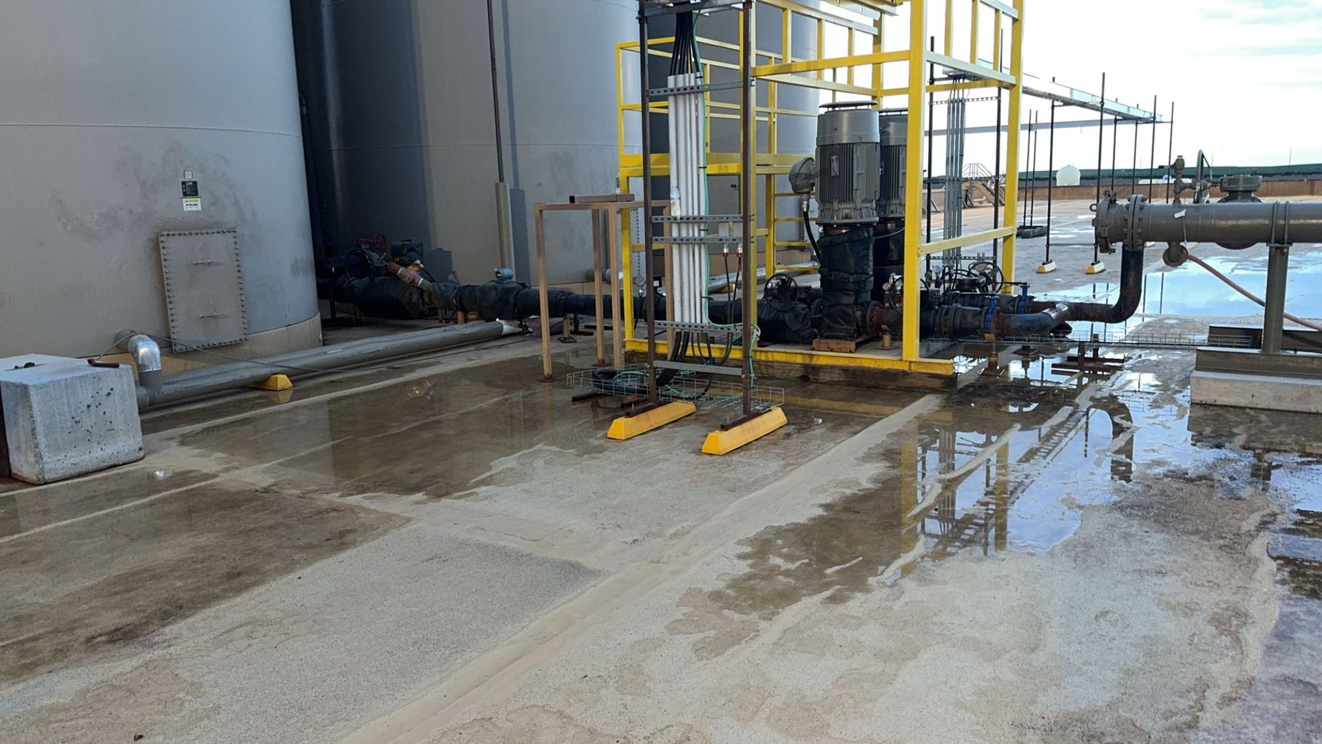 Industrial walkway with yellow railings, wet concrete, and pipes beside a building.