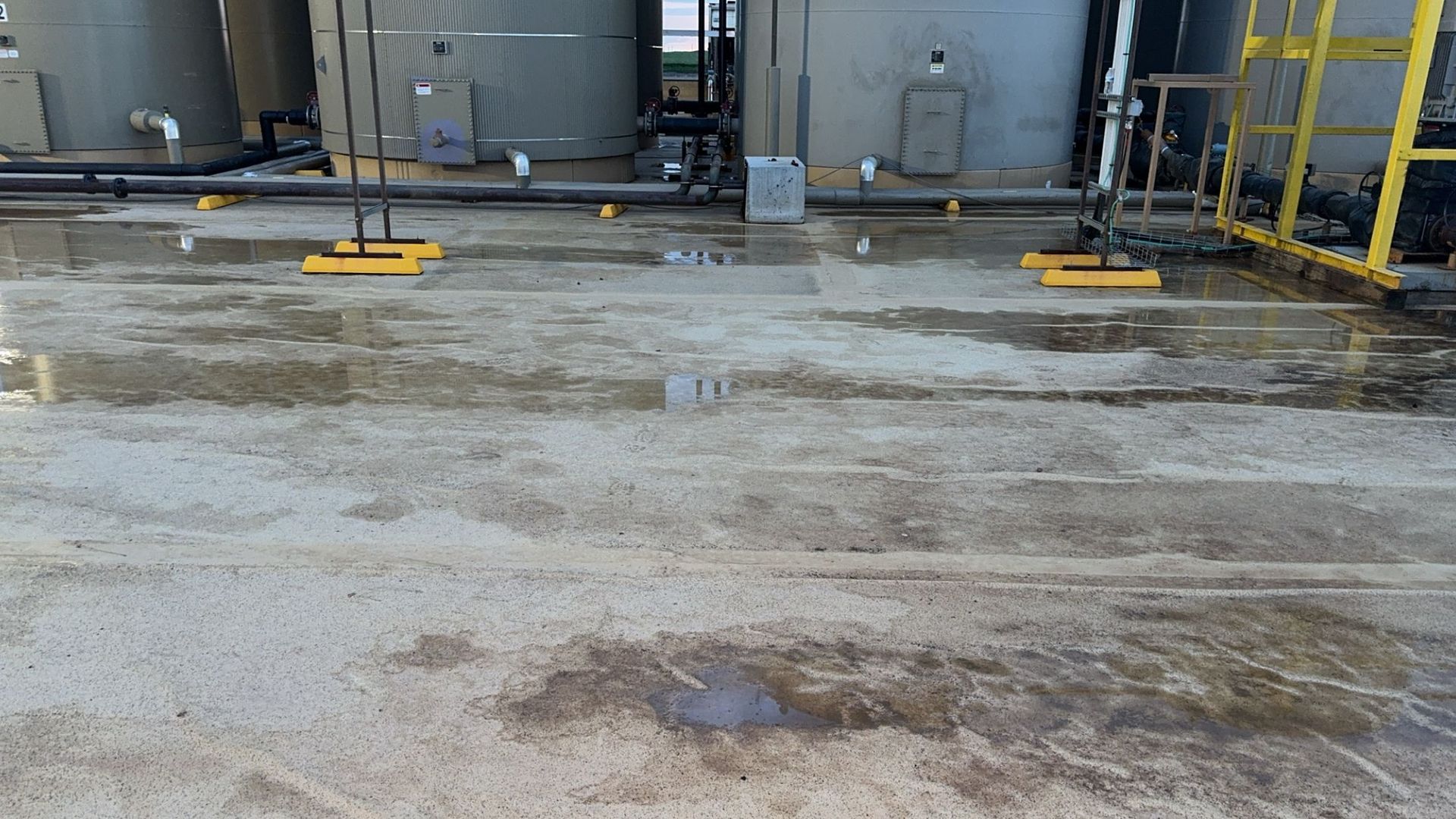 Industrial tank area with wet concrete ground, yellow railings, and metal equipment in the background