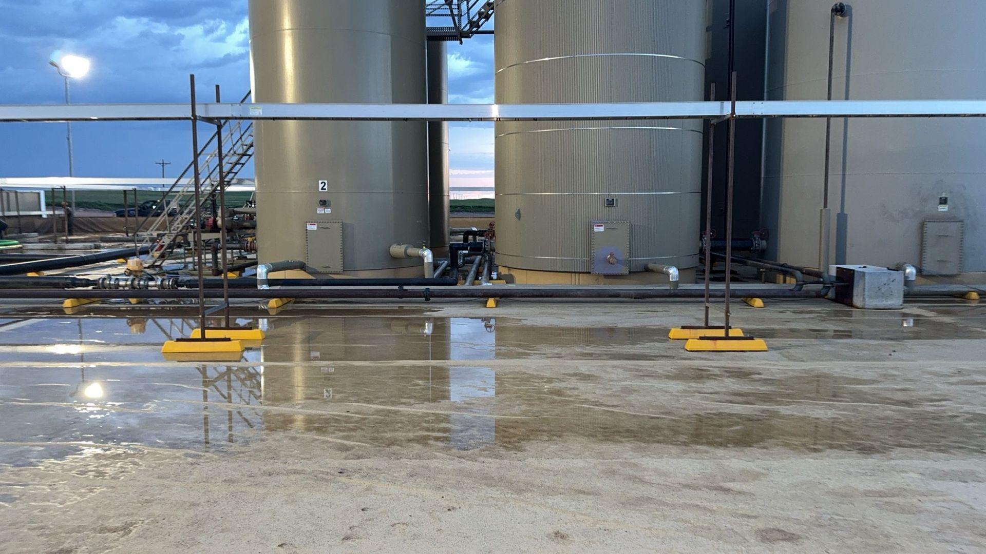 Industrial tanks and pipes in a wet refinery yard at dusk, with puddles reflecting the structures