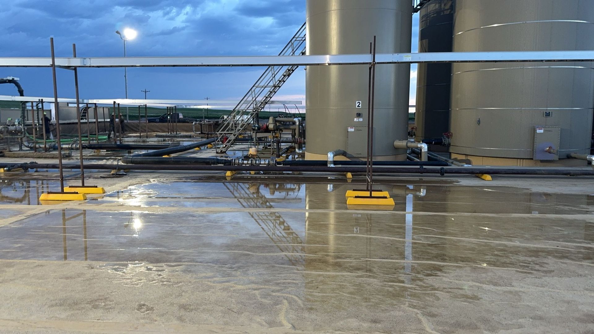 Wet industrial yard with large storage tanks, pipes, and a cloudy sky reflected on the ground