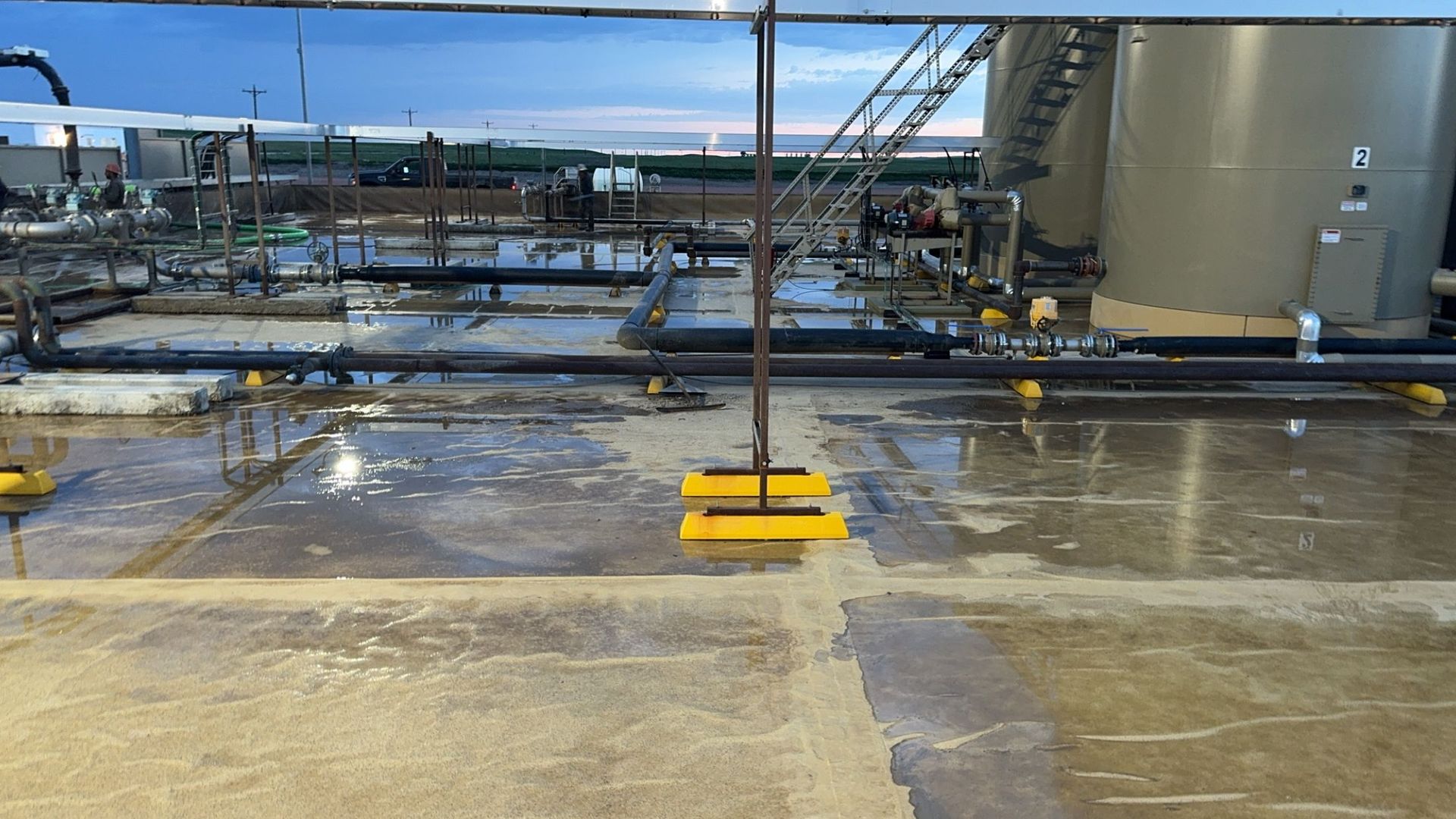 Industrial rooftop with wet concrete, pipes, tanks, and a yellow safety barrier.