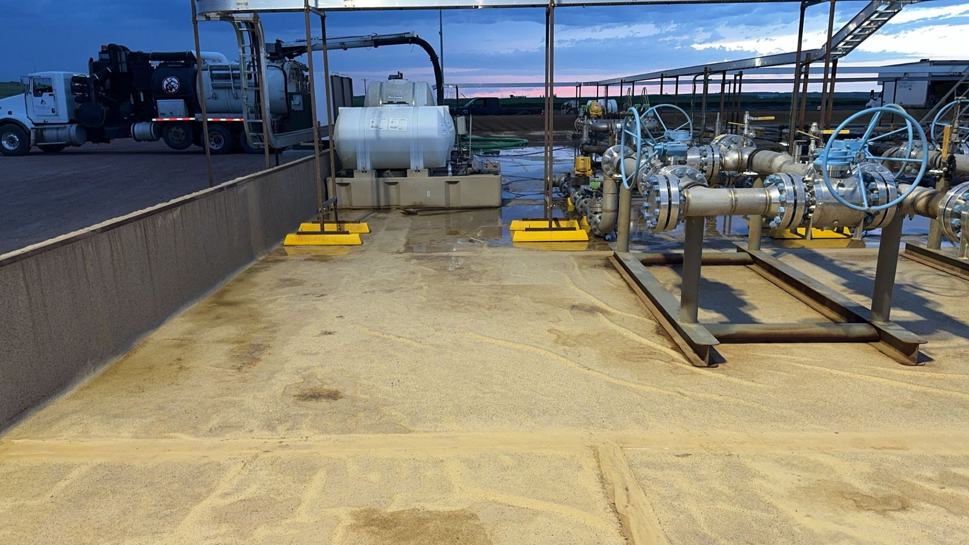Industrial pipeline and tank equipment on a concrete pad at dusk, with yellow safety markings.
