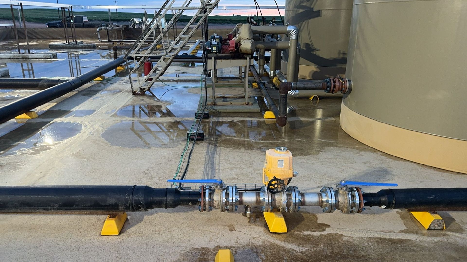 Industrial pipes and valves on a wet concrete platform beside a large storage tank.