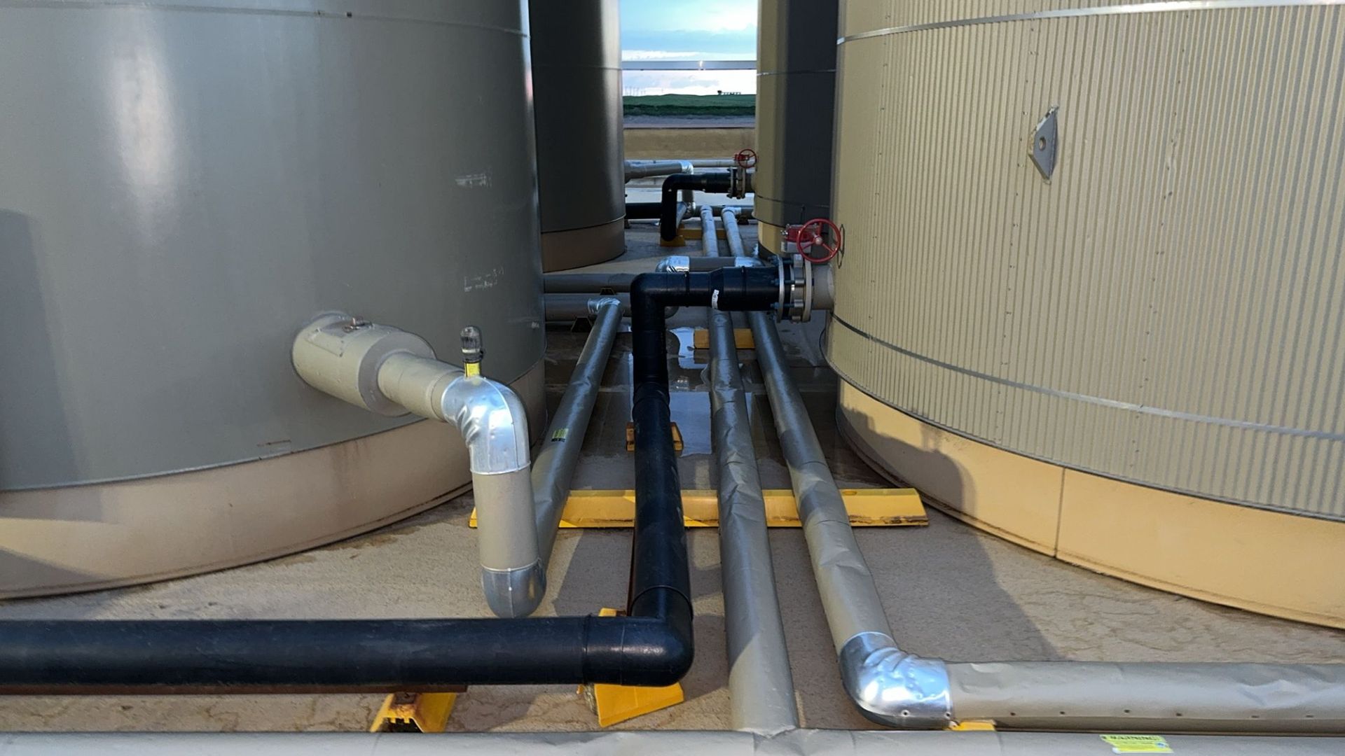 Industrial pipes between large tanks on a rooftop, with black and silver conduits running along the ground.