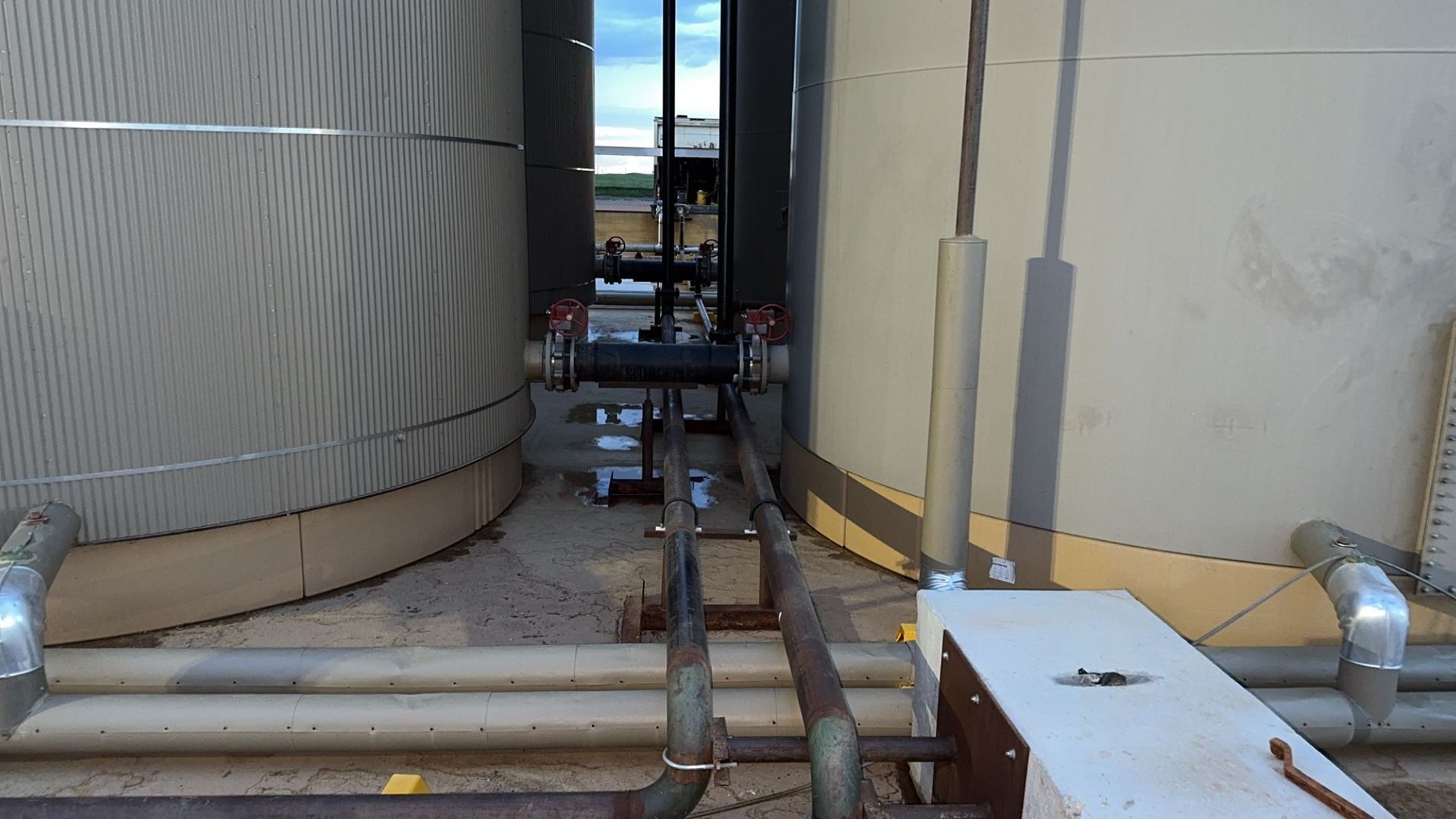 Narrow industrial walkway between large tanks with exposed pipes and debris on the floor