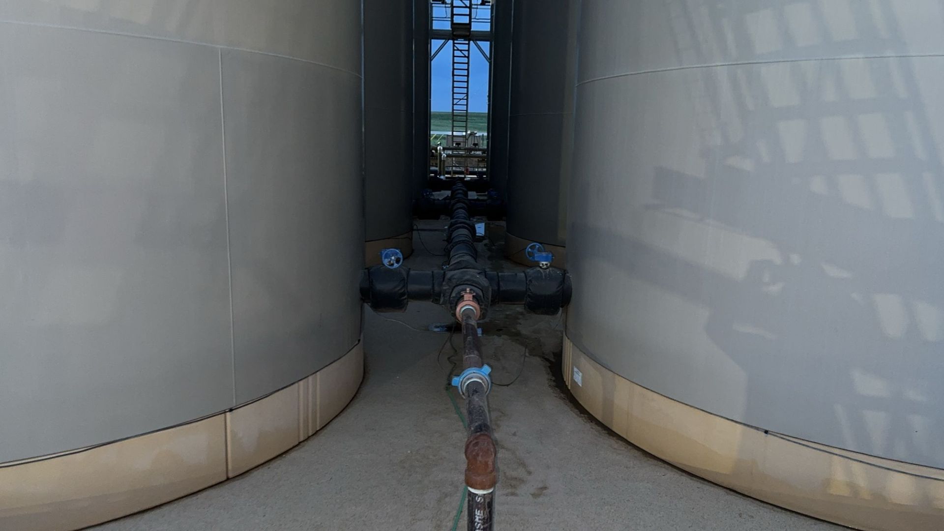 Two large industrial tanks with a narrow walkway and pipework between them under bright daylight