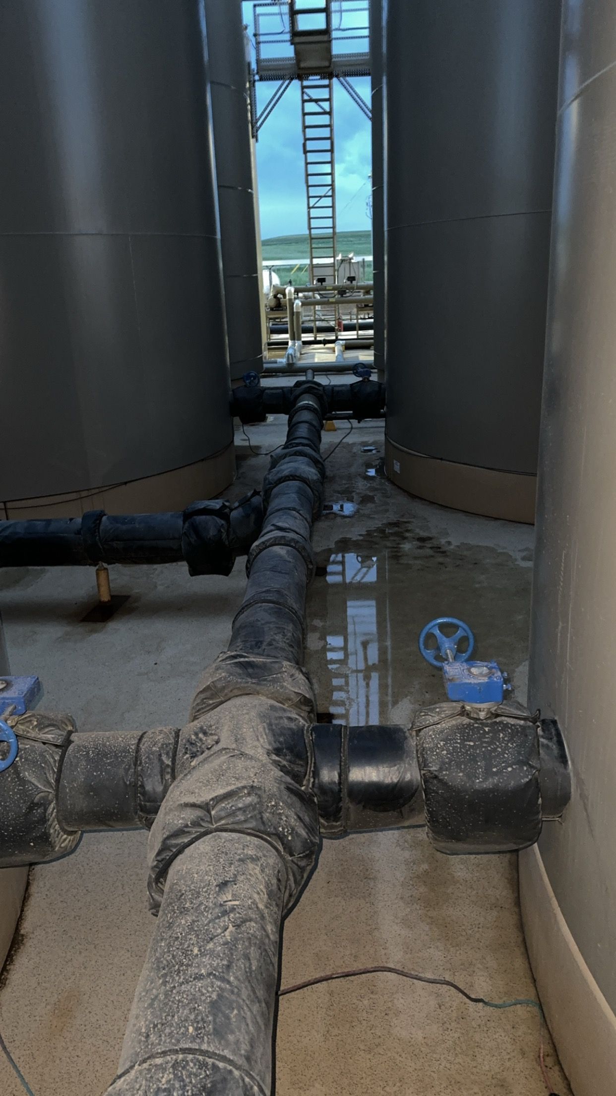 Industrial tank area with large pipes and valves between metal silos, viewed down a narrow walkway.