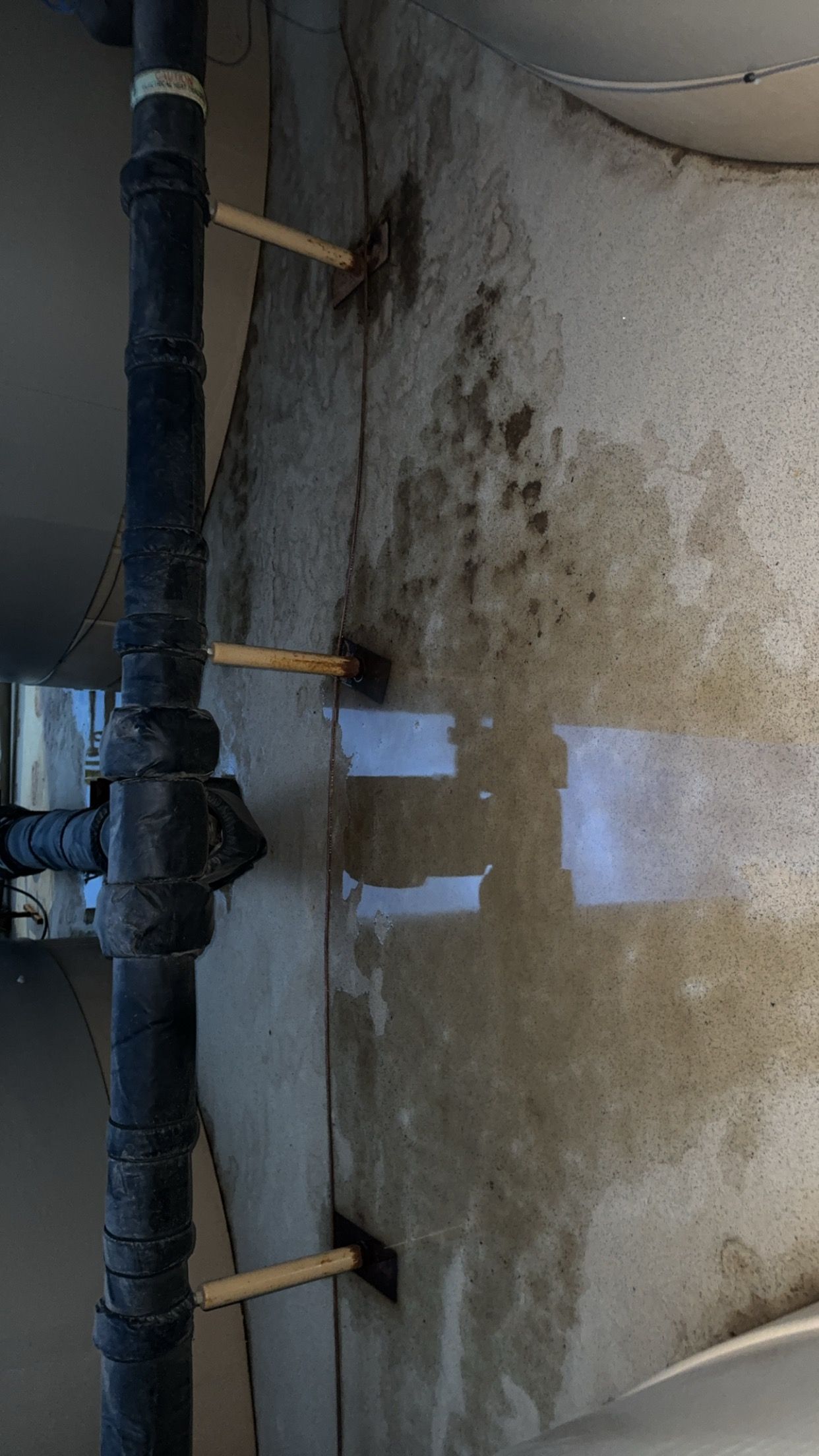 A black pipe beside a stained concrete wall with a small water puddle on the floor.