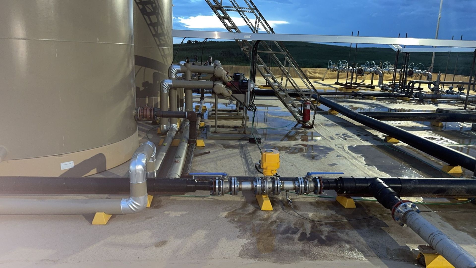 Industrial facility with large tan tank, pipes, and yellow railings in a metal structure under bright daylight