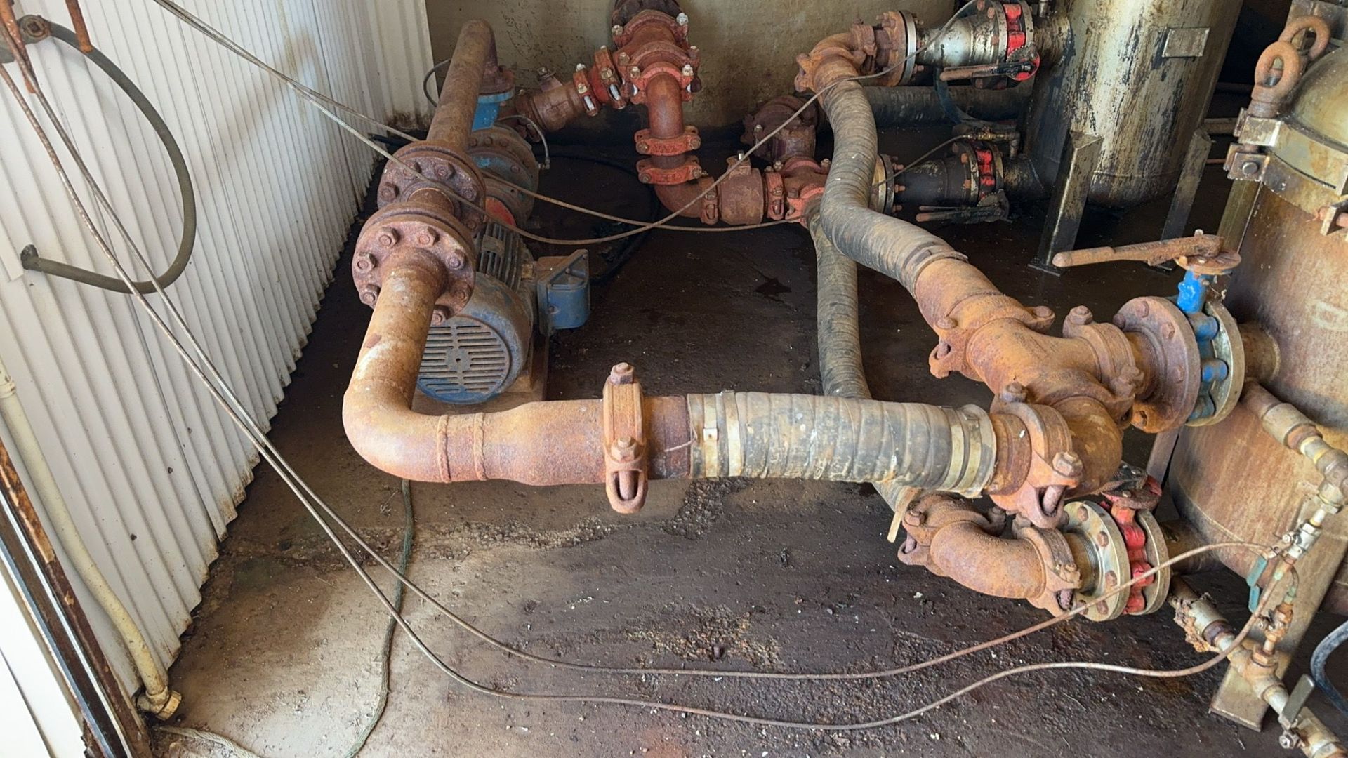 Rusty industrial pipes and valves in a cramped utility area, with white wall and floor grate.