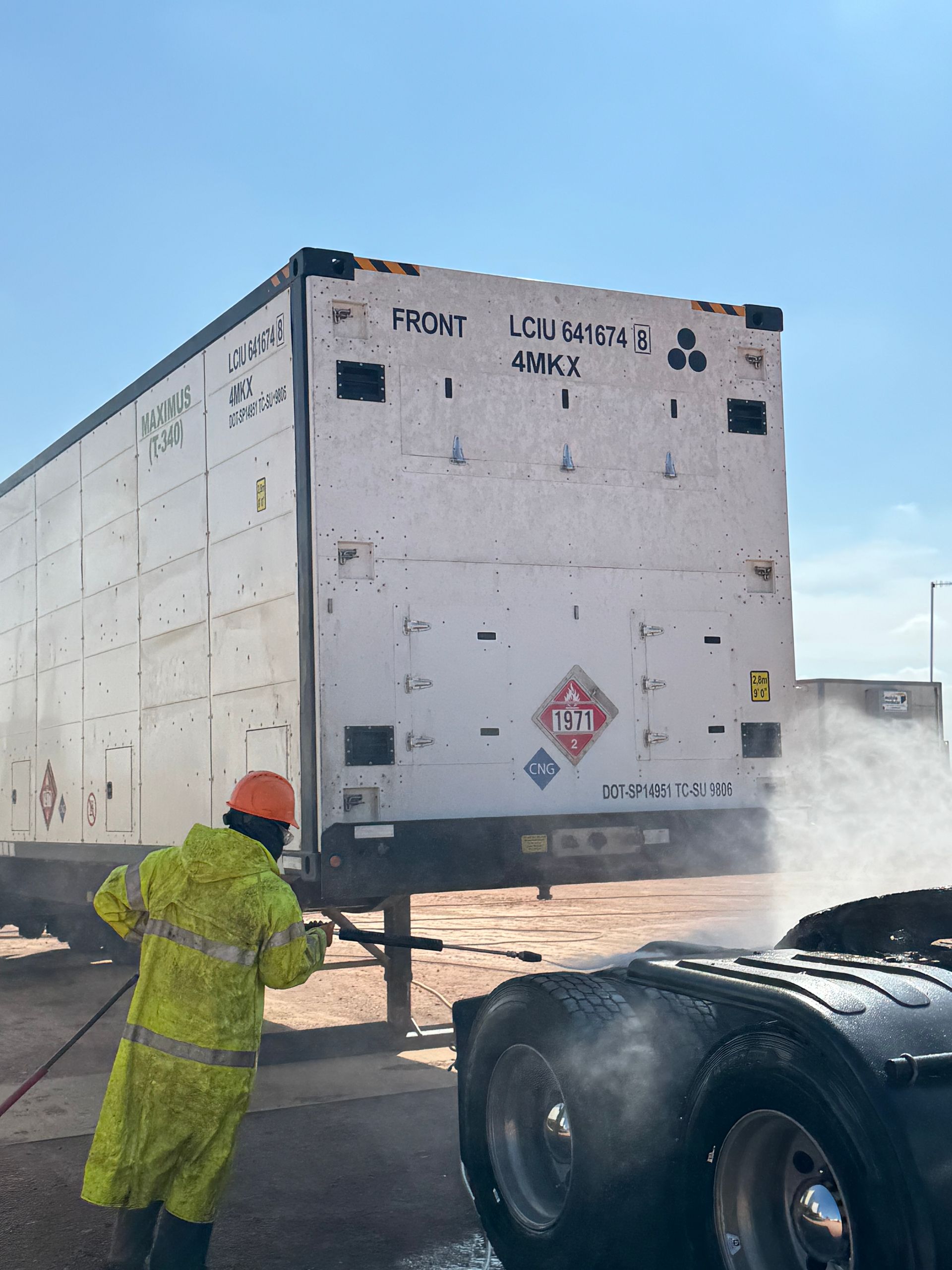Worker in yellow safety gear inspects a white trailer with steam or spray near its wheels.