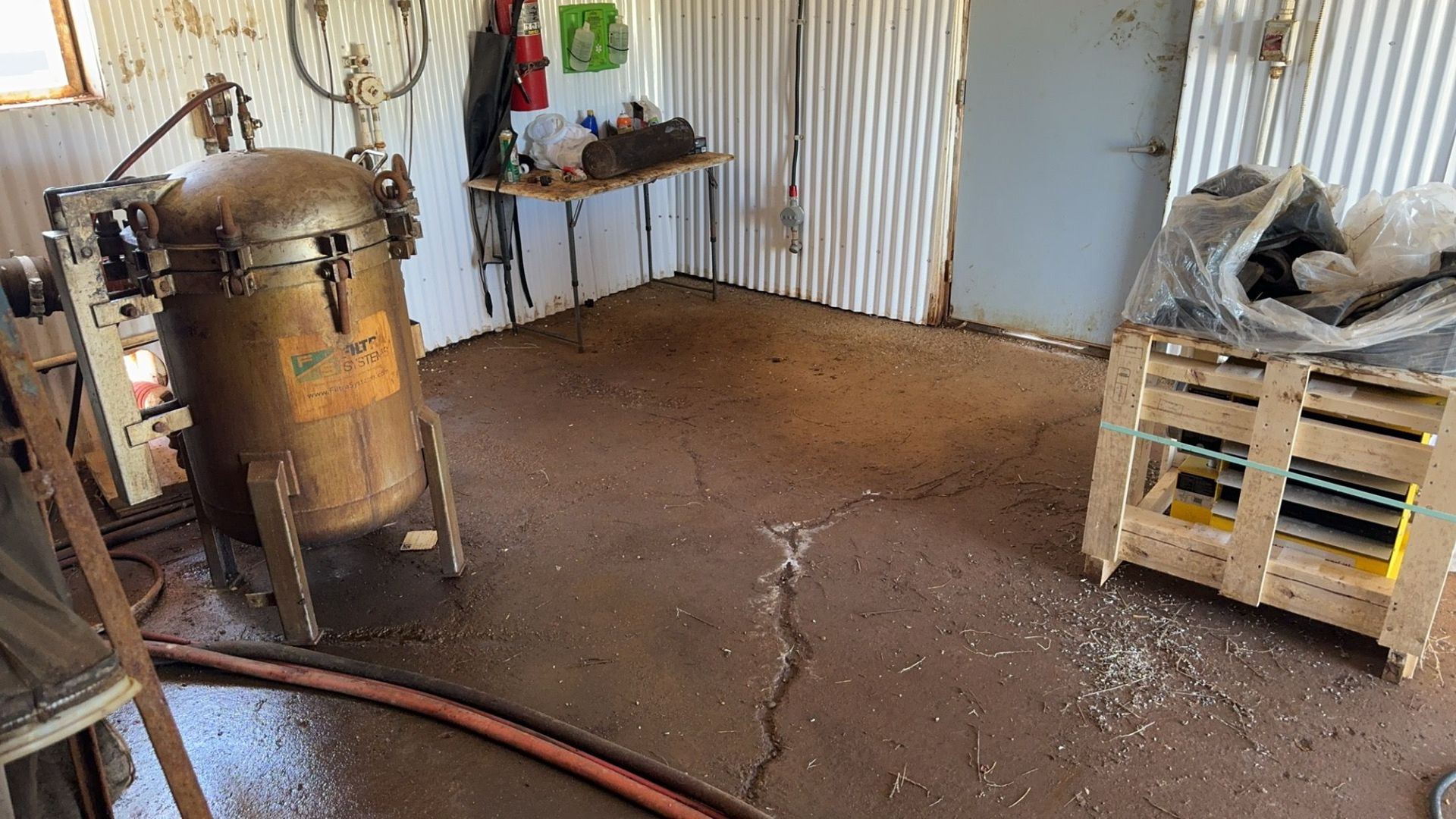 Workshop with air compressor, coiled hose, and pallet stack on a dirt floor in front of a gray door.