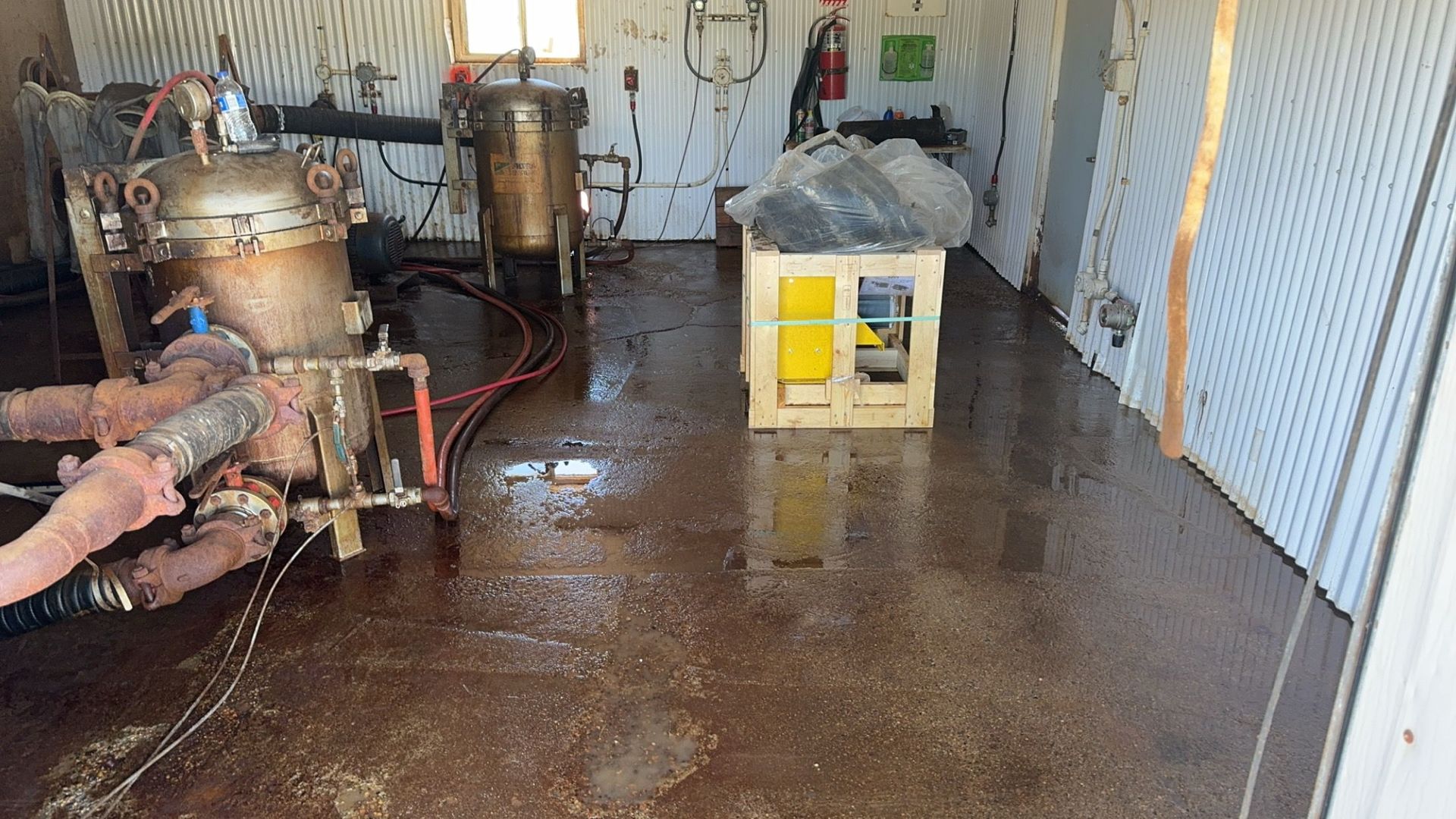 Industrial room with wet concrete floor, tanks and pipes along the wall, and a yellow machine near the center.