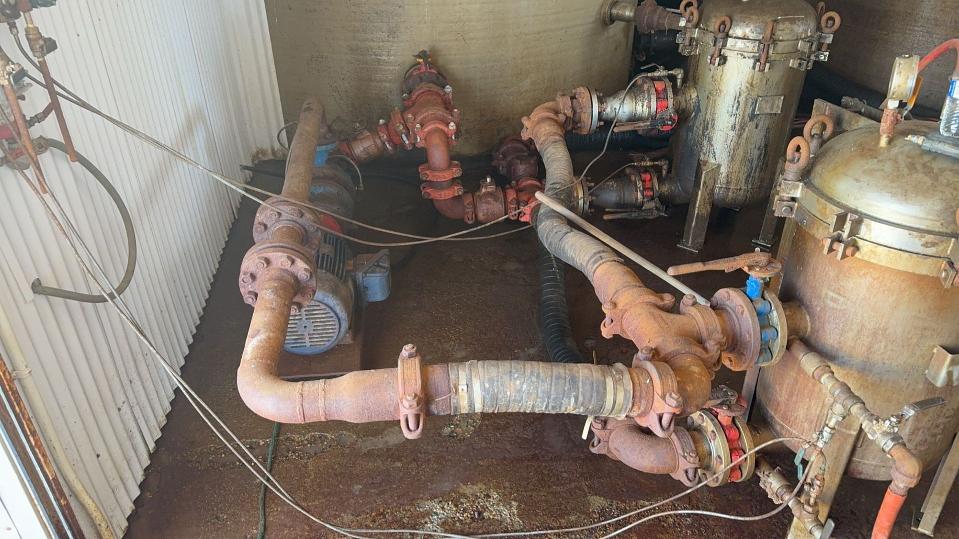 Industrial pipes and valves in a mechanical room, with rusted metal equipment and a large tank.