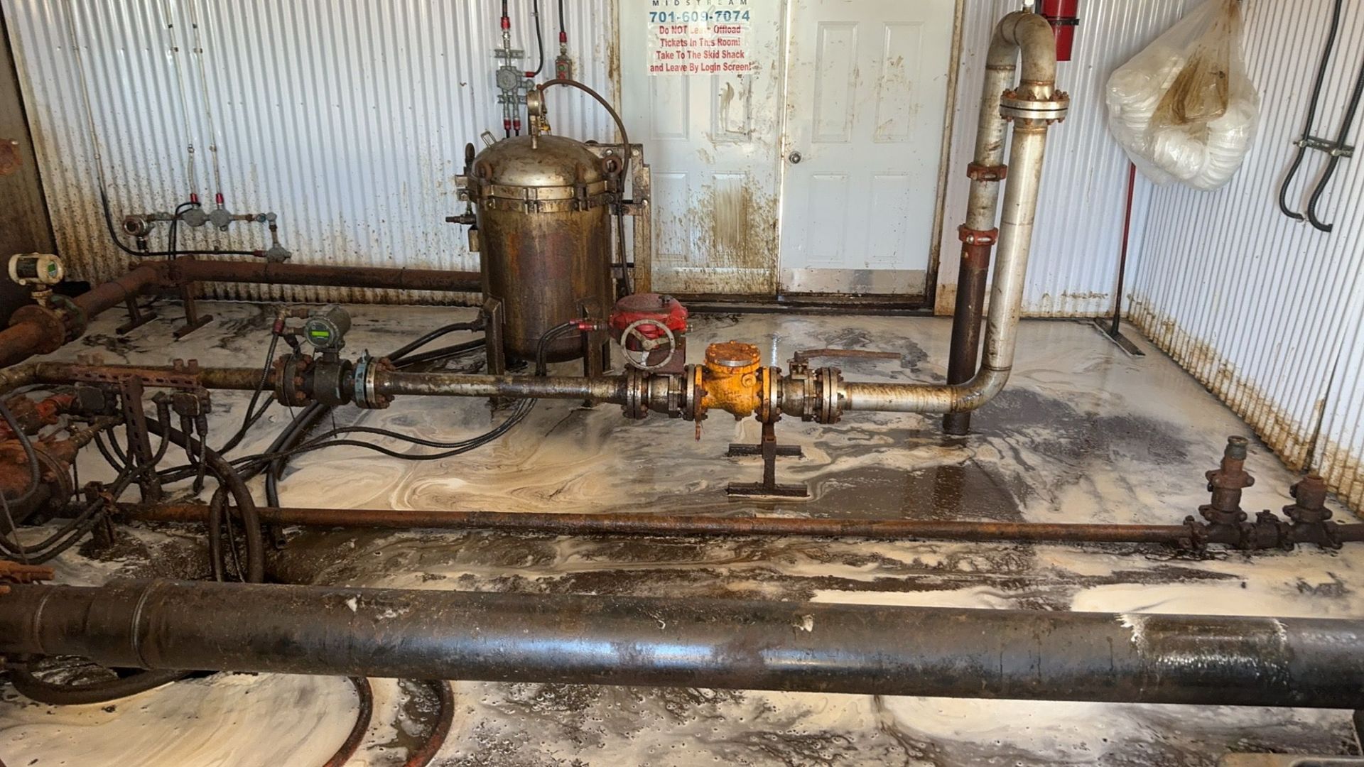 Industrial boiler room with corroded pipes, a central tank, and a small gas flame on the floor.