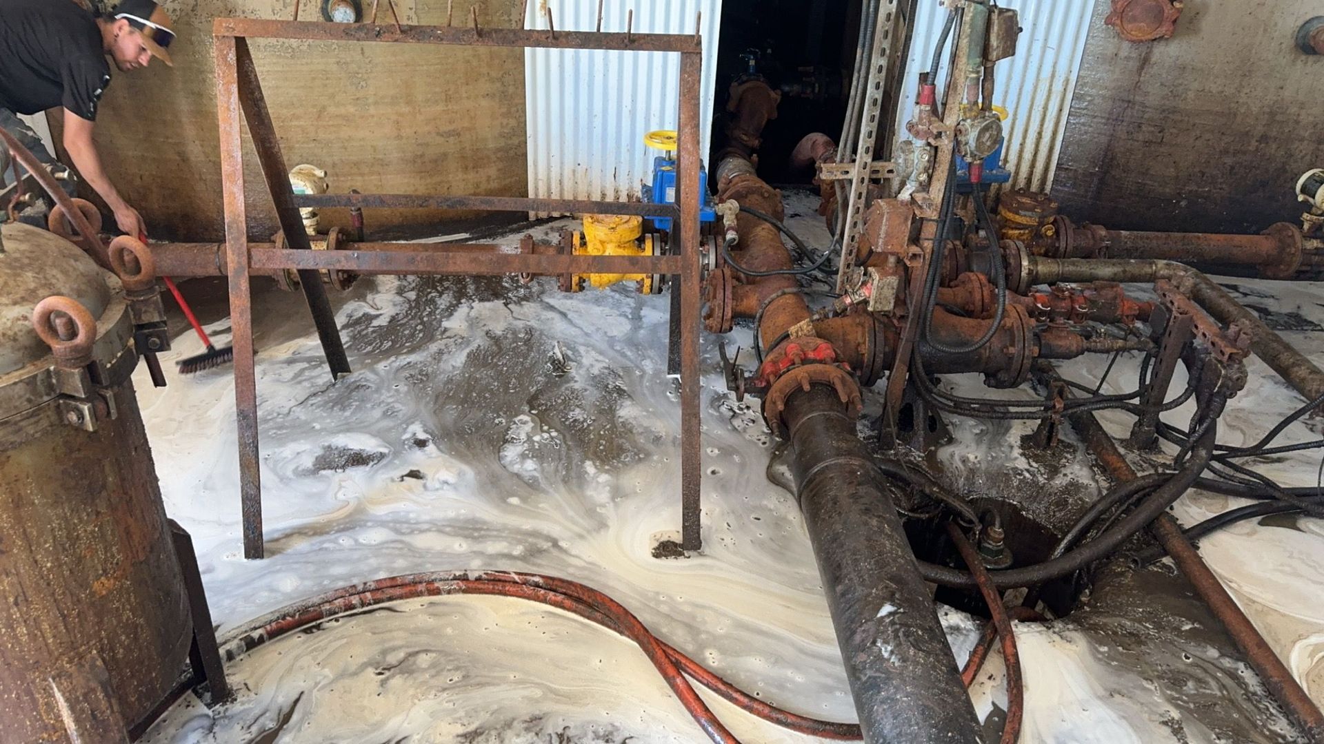 Industrial machinery with rusted pipes and hoses leaking onto a wet concrete floor in a factory area