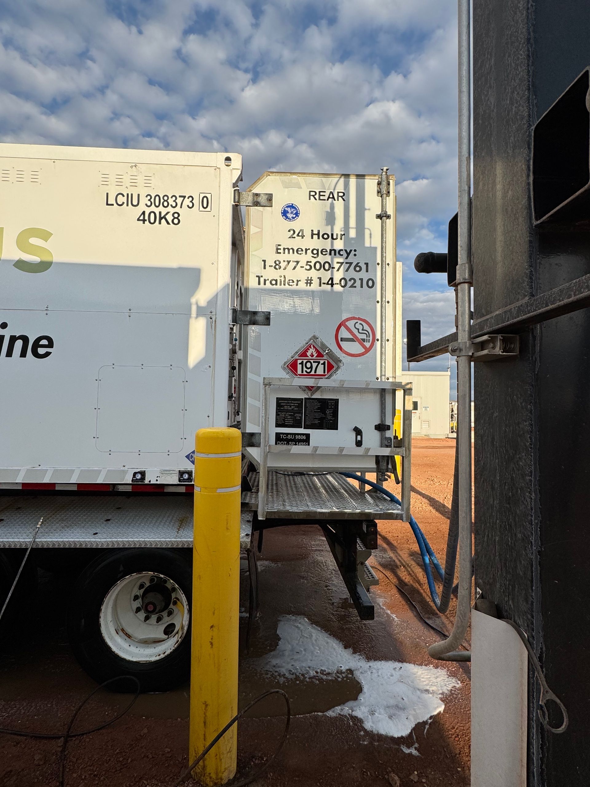 White refrigerated truck at a loading dock with yellow bollard and wet ground under cloudy sky