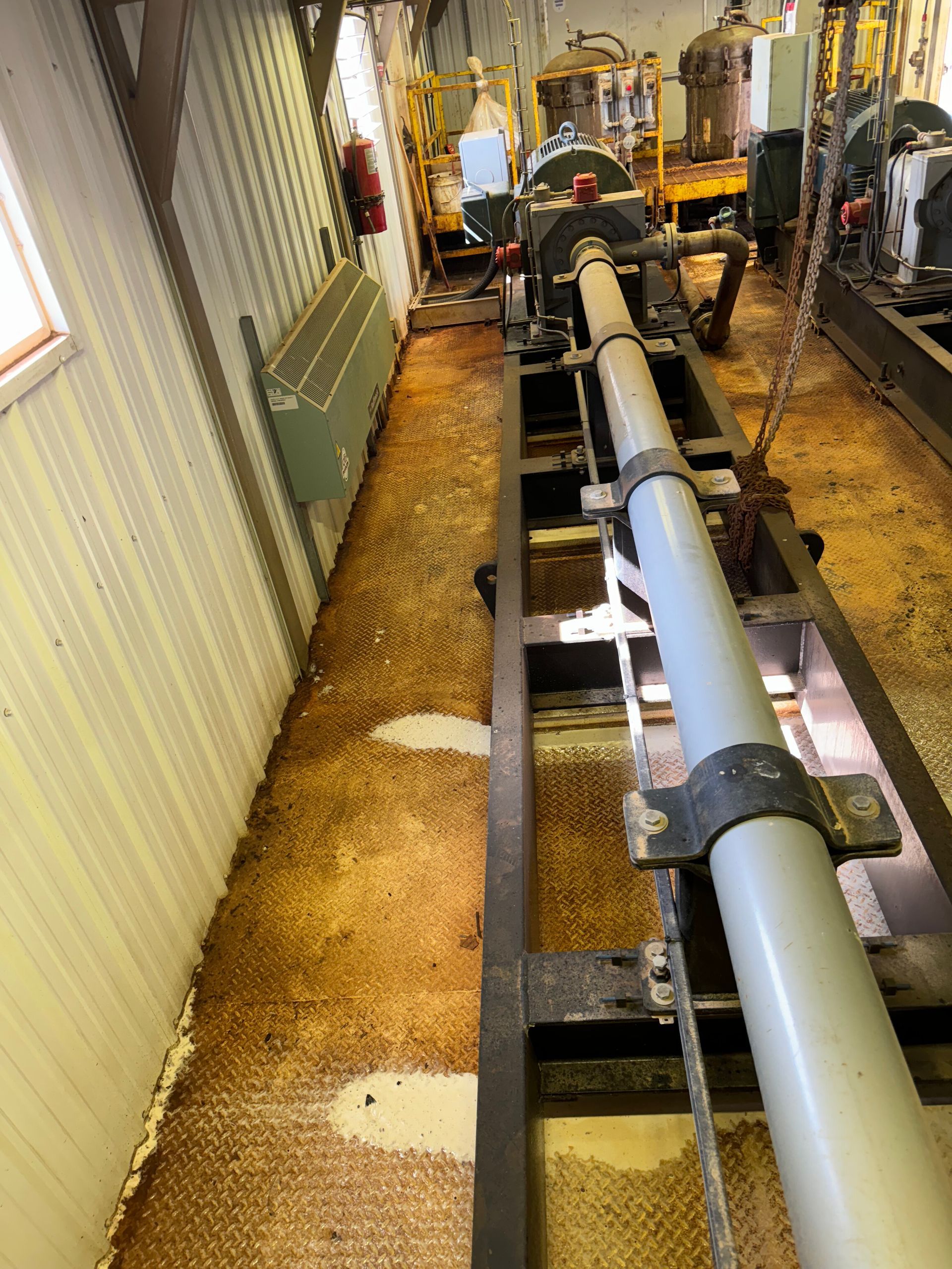 Interior of an industrial facility with large pipes running through a narrow corridor along corrugated metal walls.