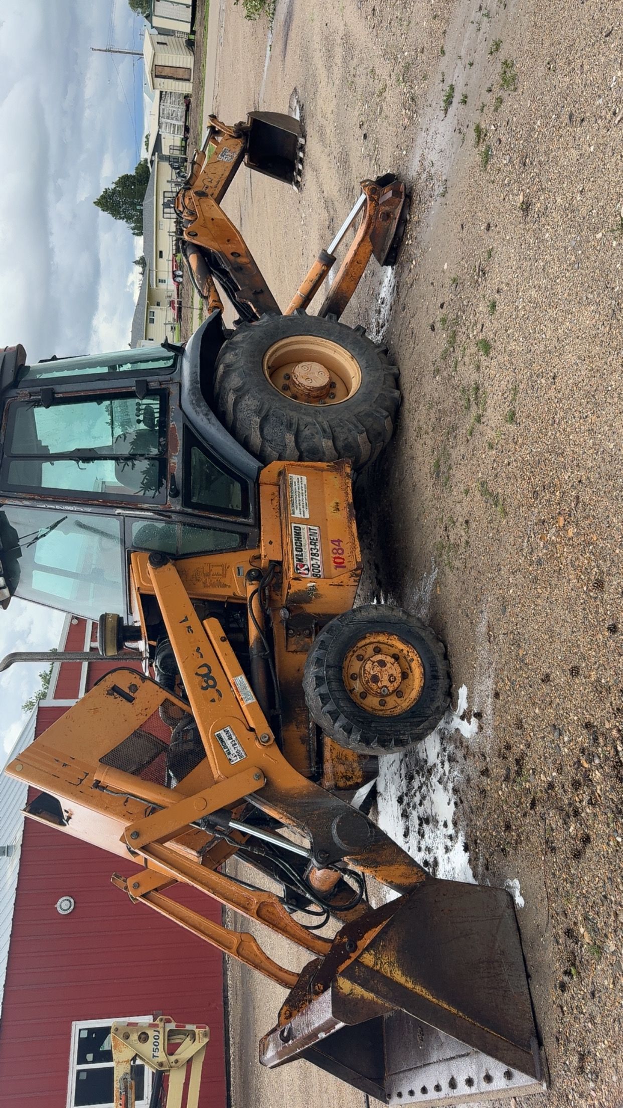 Orange backhoe loader parked in a snowy yard beside a red building and a chain-link fence