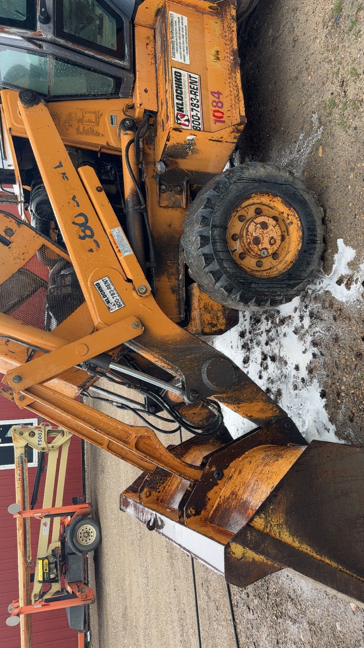 Orange excavator clearing snow beside a muddy roadside, with its bucket raised.