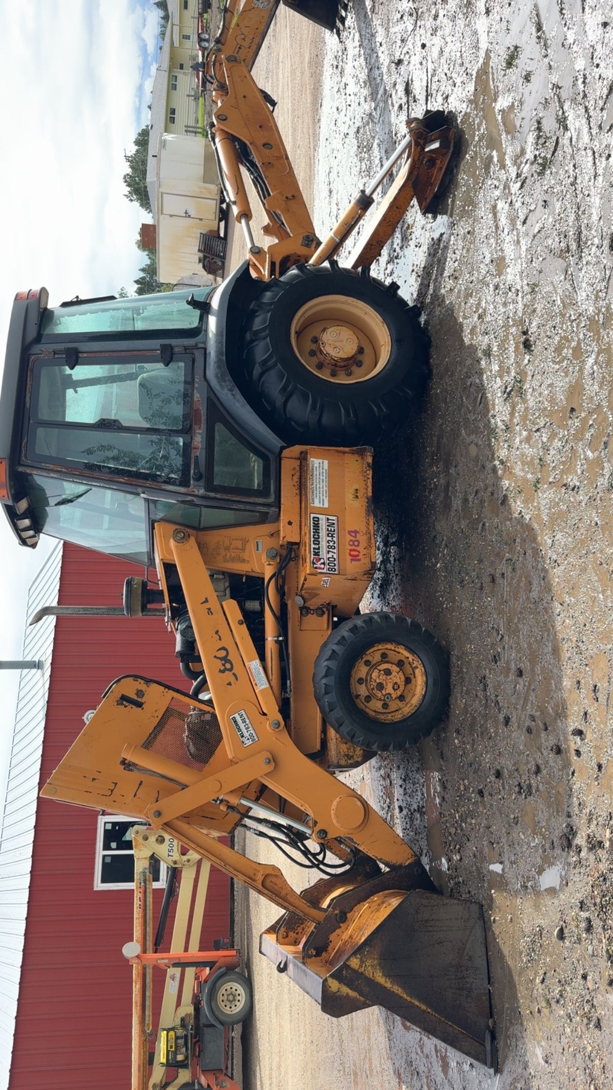 Orange backhoe loader on a snowy lot beside a red building, with its bucket raised and digging arm extended