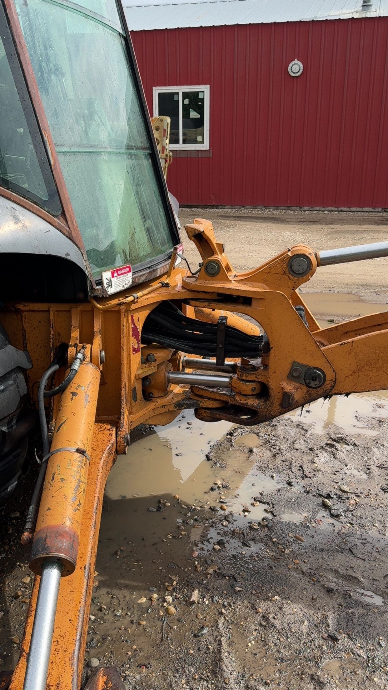 Close-up of a yellow excavator arm and cab beside a red building on muddy ground.