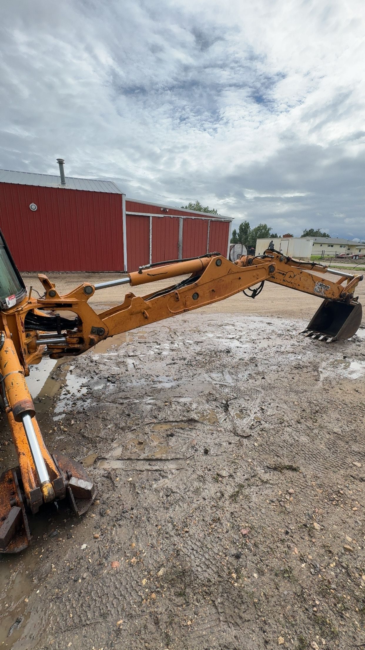 Yellow excavators beside a red building on a muddy construction site under a cloudy sky
