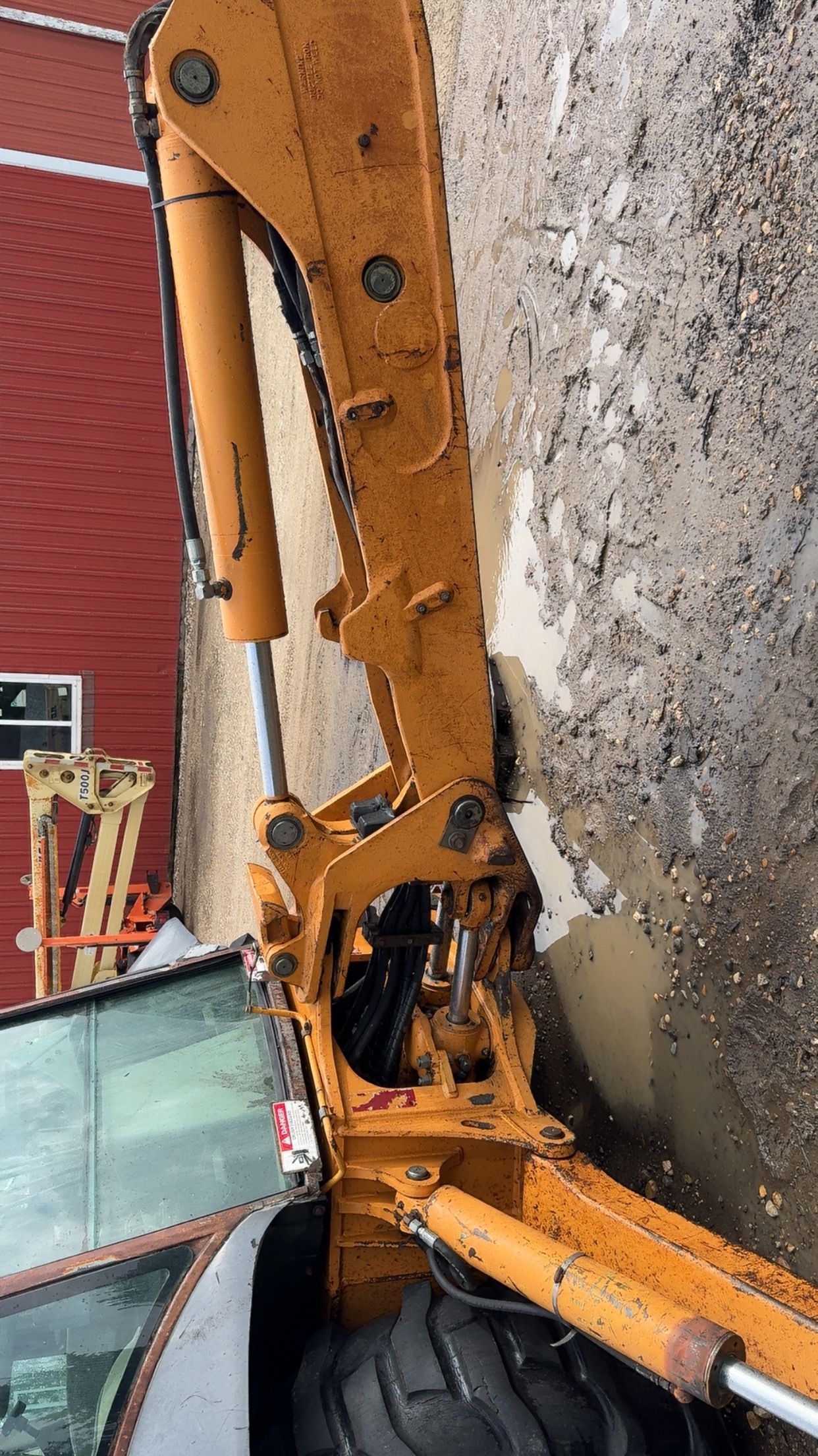 Orange excavator arm beside a wet gravel surface near a red building