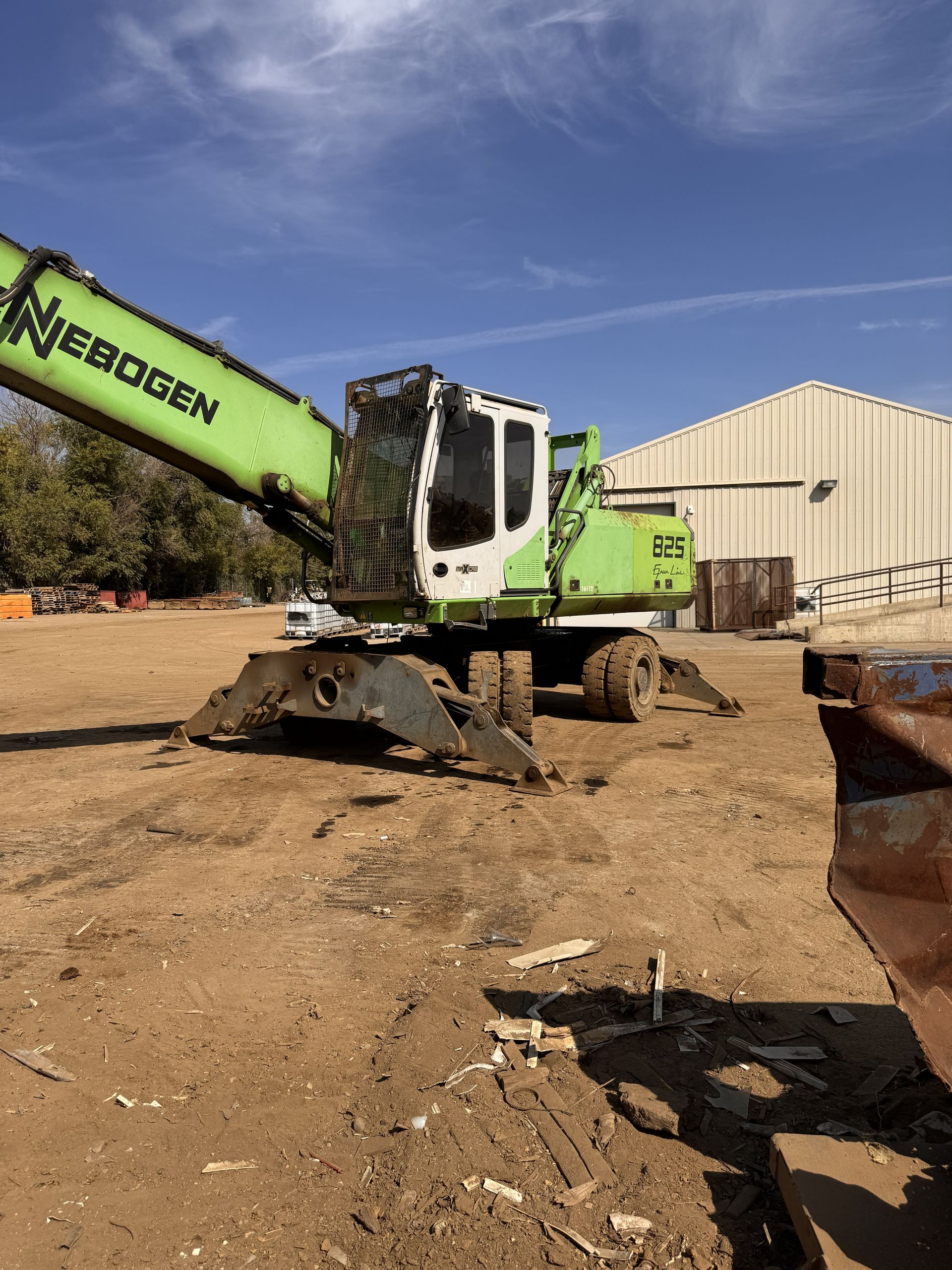 Green excavator at a construction site beside a beige warehouse under a clear blue sky