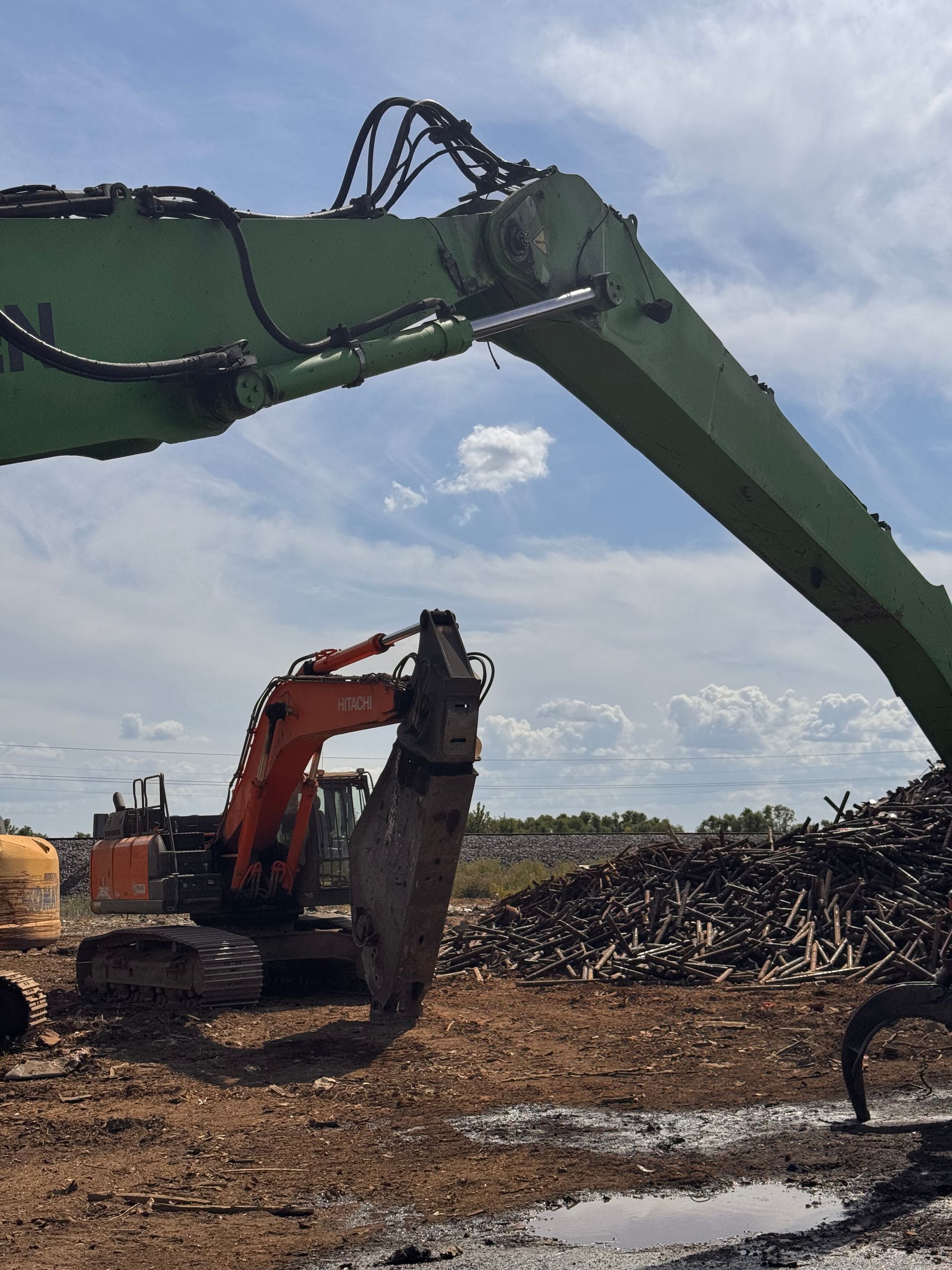 Green excavator and orange excavator working at a scrap metal yard under a cloudy sky